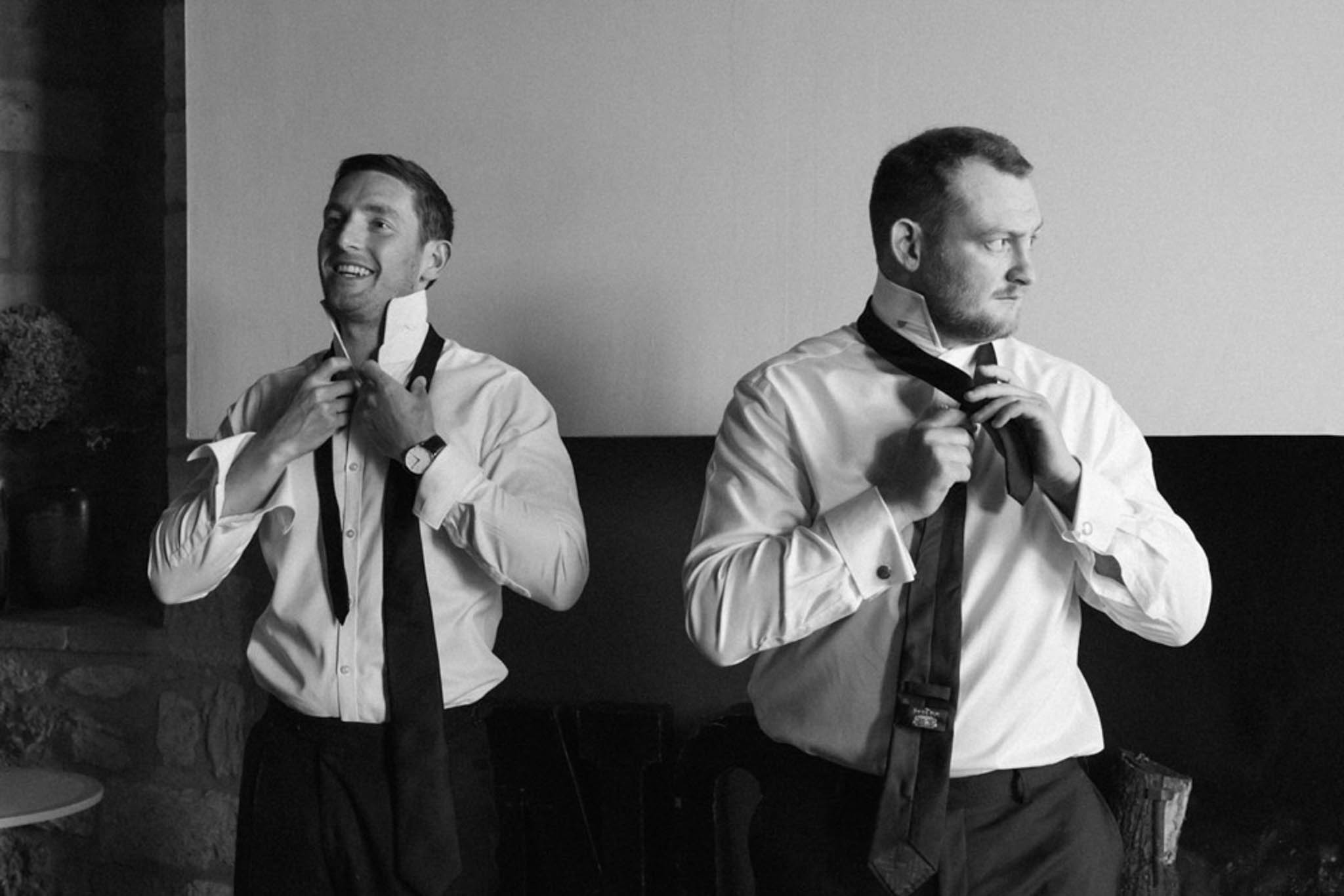 Black and white two-shot of groom and groomsman adjusting dark ties in white shirts with suspenders