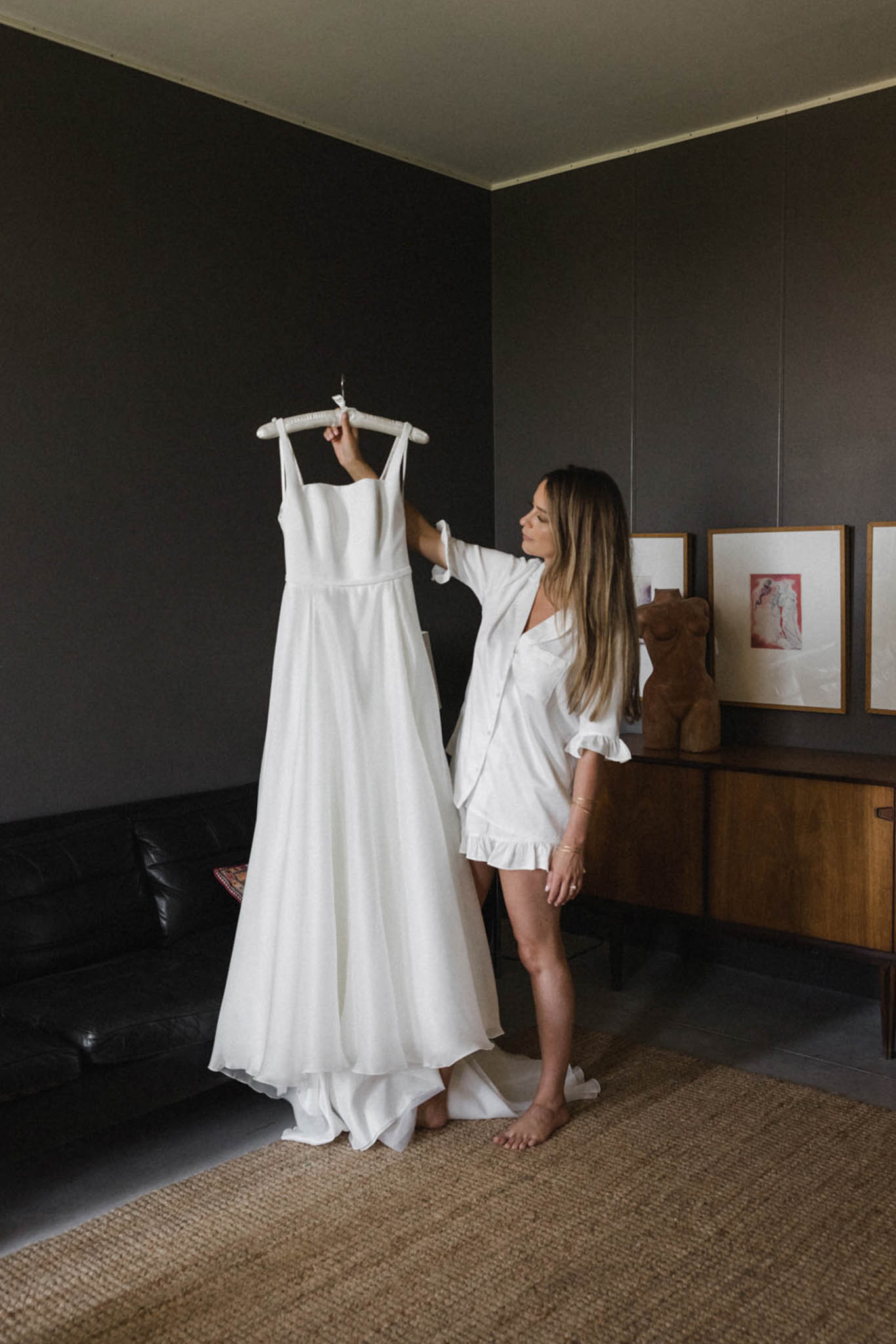 A getting-ready moment showing the bride standing barefoot in an indoor room, holding up her wedding dress on a white padded hanger and looking at it. She is wearing a white satin short robe with ruffled trim and has long blonde highlighted hair worn loose. The wedding dress is ivory with thin square-neck straps, a fitted bodice, and a full A-line skirt that pools on the floor. The room has dark charcoal-painted walls, a black leather sofa to the left, a natural woven jute rug on the floor, and a mid-century style wooden sideboard in the background topped with a sculptural decorative object. Three framed artworks with warm wood frames hang on the wall behind her, including one with a pink figurative print. The overall interior styling is modern and moody. The shot is a full-length portrait taken from a slight distance, capturing both the bride and the full length of the dress.