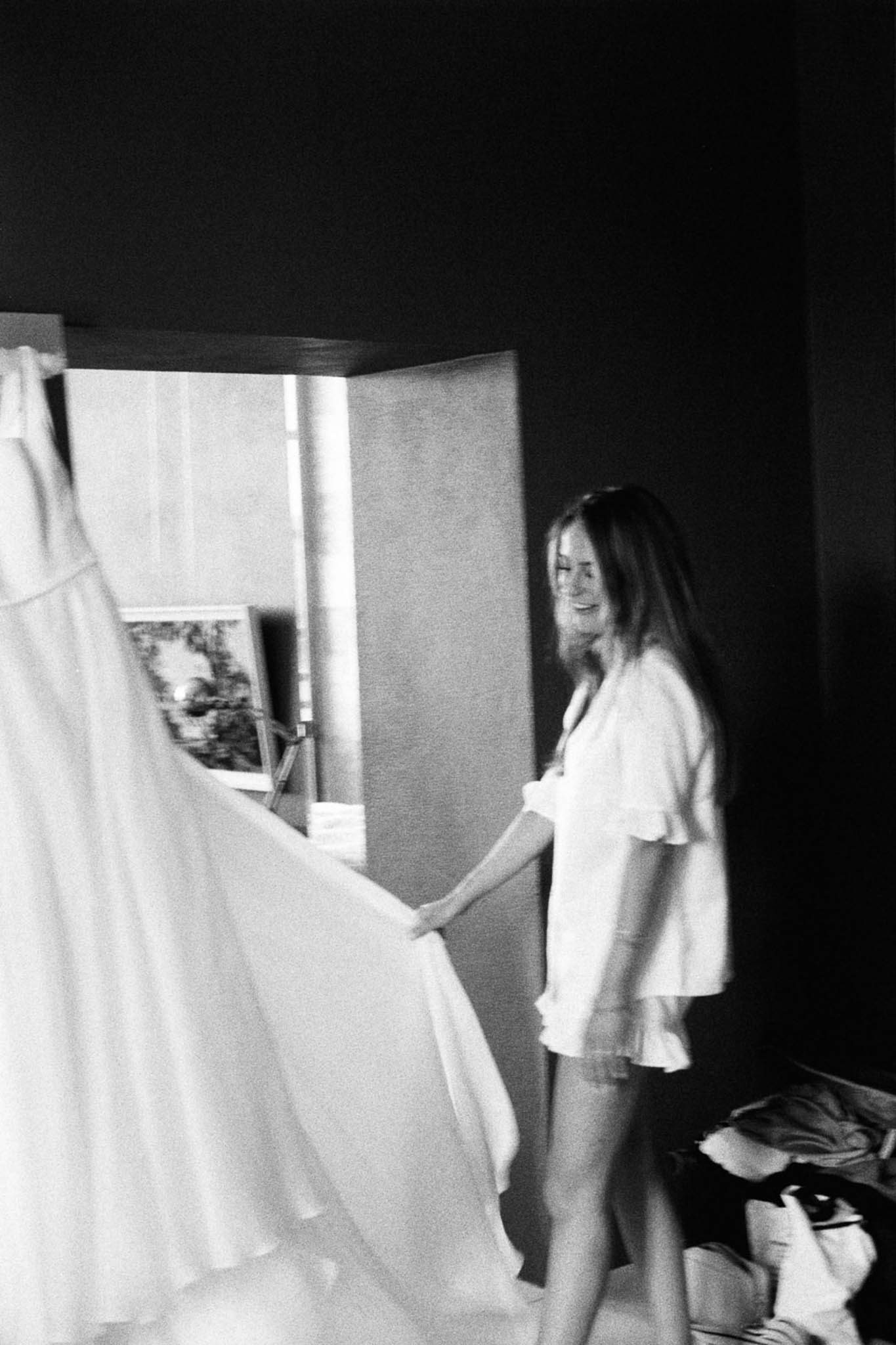 A black-and-white getting-ready shot of a woman, likely the bride, standing in a darkened indoor room and handling what appears to be a large, full-skirted wedding gown hanging to her left. She is wearing a short-sleeve ruffled pajama set and has long straight hair, and she is smiling as she touches the dress fabric. A framed picture and a folding screen or room divider are visible in the background, with natural light coming through a narrow window, and bags or belongings are visible on the floor to the right. The image has a grainy, high-contrast film aesthetic with deep dark tones in the background and bright highlights on the dress and figure. The shot is slightly soft in focus, giving it a candid, documentary feel.