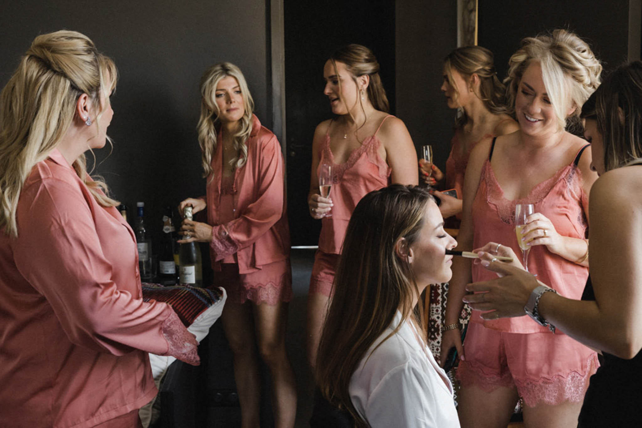 A getting-ready scene shot indoors in a darkly decorated room, showing approximately six women gathered together. The bride, identifiable by her white robe, is seated and having makeup applied, while surrounded by bridesmaids or members of the bridal party wearing matching coral-pink satin lace-trimmed camisole and shorts sets, along with one woman in a matching coral-pink satin robe. Several women are holding champagne flutes, and one woman on the left is opening a bottle of wine or champagne near a small drinks setup visible in the background. Hair and makeup are in various stages of completion, with several women wearing updos and half-up styles. The image is a candid mid-shot capturing the relaxed, social atmosphere of the bridal getting-ready moment.