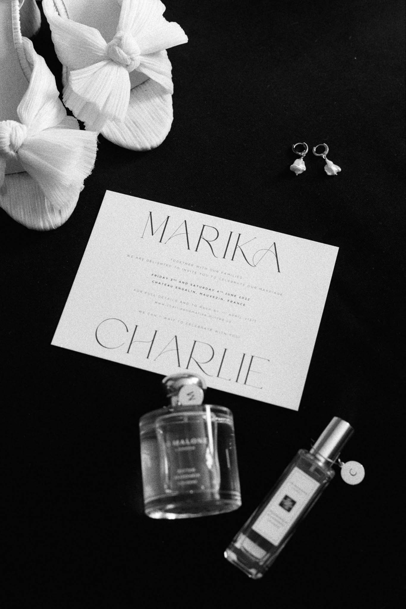 Black and white flat lay of bridal details with wedding invitation, bow sandals, pearl earrings, and perfume bottles
