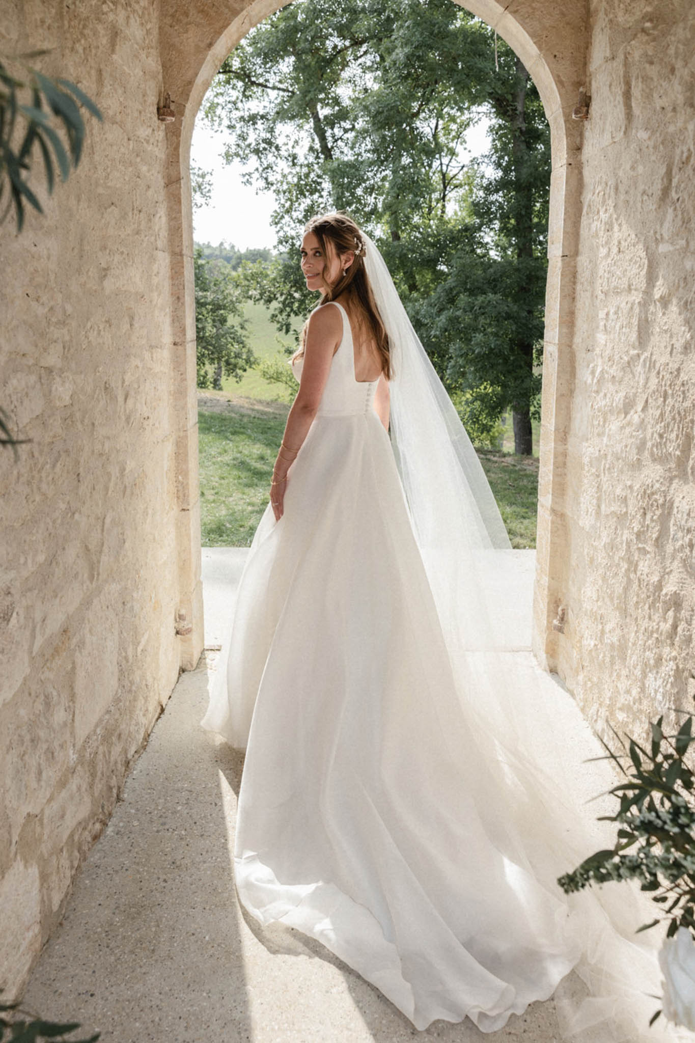 A bridal portrait taken in a stone archway passageway, likely part of a château or historic estate. The bride stands with her back to the camera and turns to look over her shoulder, smiling. She wears an ivory ball-gown style dress with a square low back, wide straps, and a full tulle skirt with a long cathedral-length veil that pools on the ground behind her. Her brown hair is worn half-down with a small floral or crystal hair accessory. The composition is a full-length portrait framed by the arched limestone doorway, with greenery and open grounds visible through the arch in the background. A small sprig of eucalyptus foliage is visible in the lower right corner. The styling is clean and classic with minimal ornamentation.