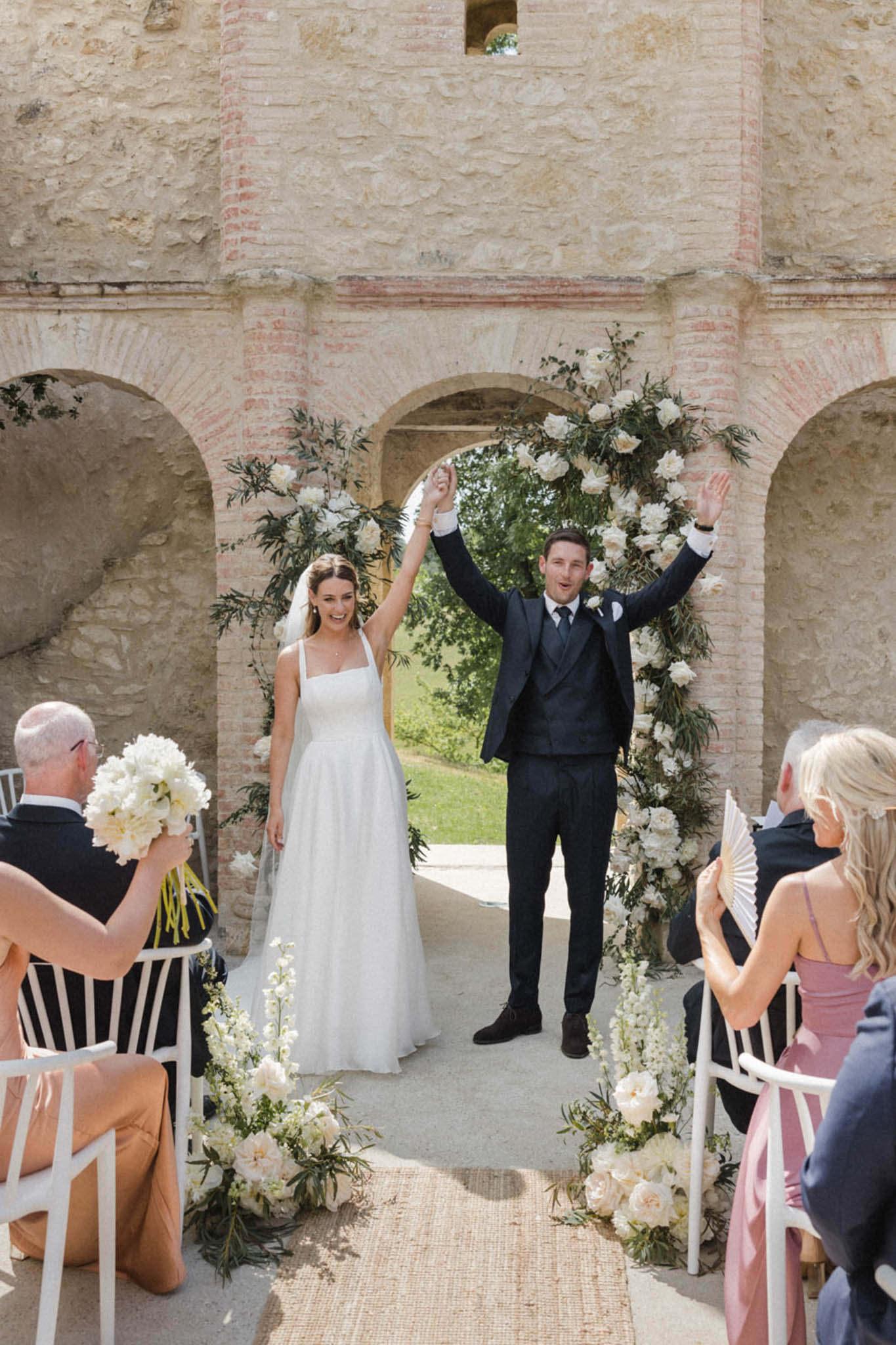 Bride and groom raise joined hands walking from circular peony arch at stone ruin ceremony with seated guests