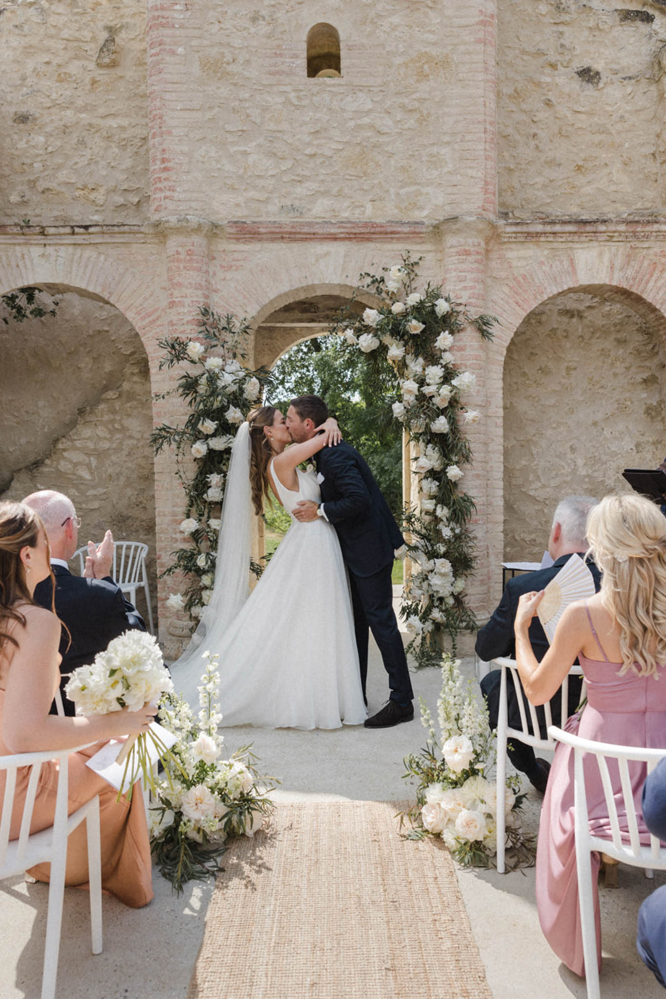 First kiss under circular ivory peony arch on jute runner before stone and red-brick arched structure