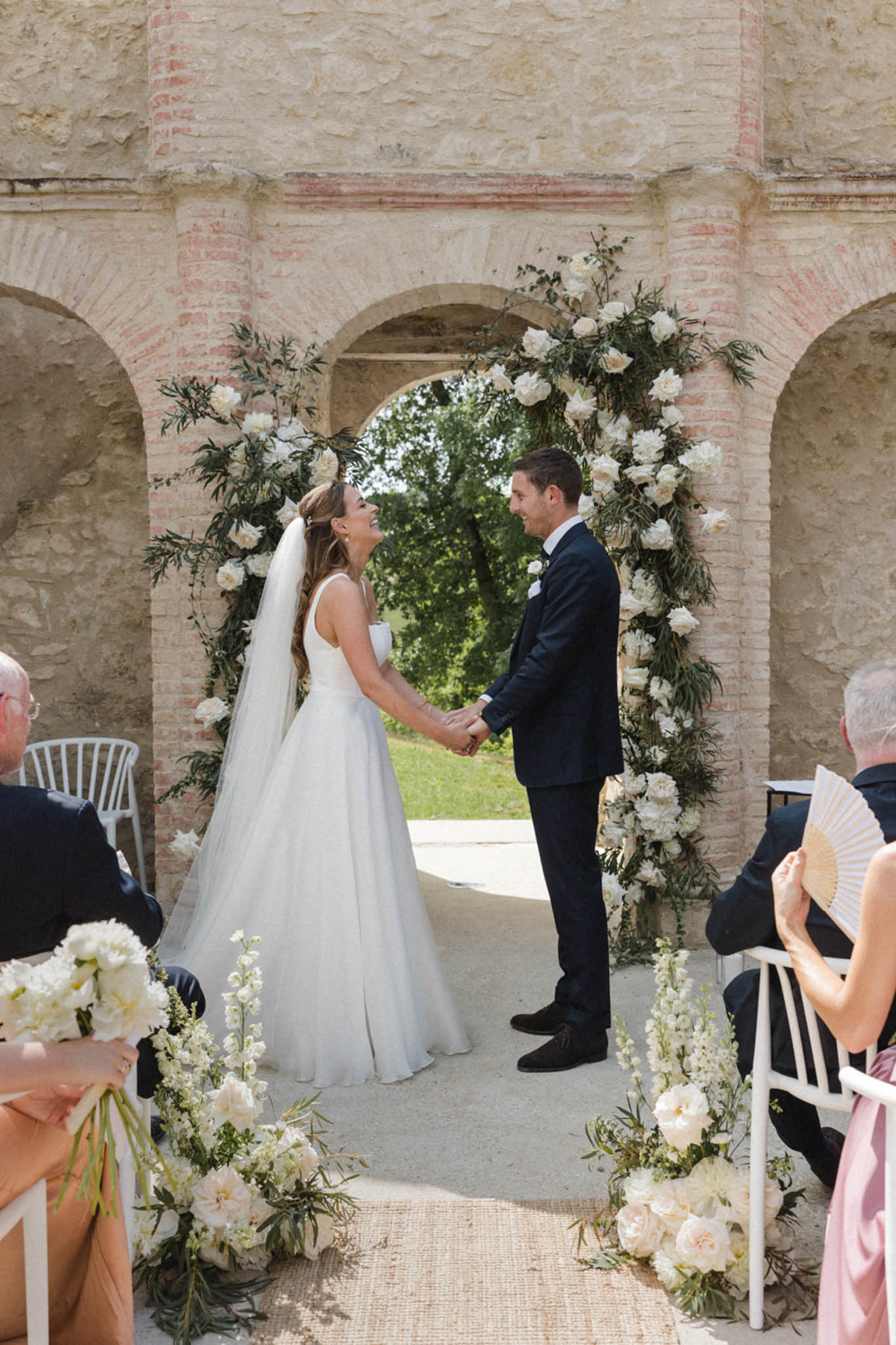 An outdoor ceremony is taking place beneath a stone arcade with brick-trimmed arches, where the bride and groom stand facing each other holding hands and smiling. The bride wears a sleeveless white ballgown with a cathedral-length veil, while the groom wears a navy suit with a white boutonniere. Behind them is a large circular floral arch filled with ivory peonies and dark green foliage, framing an open archway with a garden view beyond. The aisle is lined with clusters of ivory peonies, white delphiniums, and greenery at the base, and a natural jute runner leads toward the altar. Seated guests are partially visible on both sides, including one holding a white handheld fan and another holding a white bouquet of ivory blooms; one guest in a blush pink dress is visible to the right. The overall decor palette is ivory and green with a classic, clean aesthetic. Medium wide shot taken from the guest perspective down the aisle.
