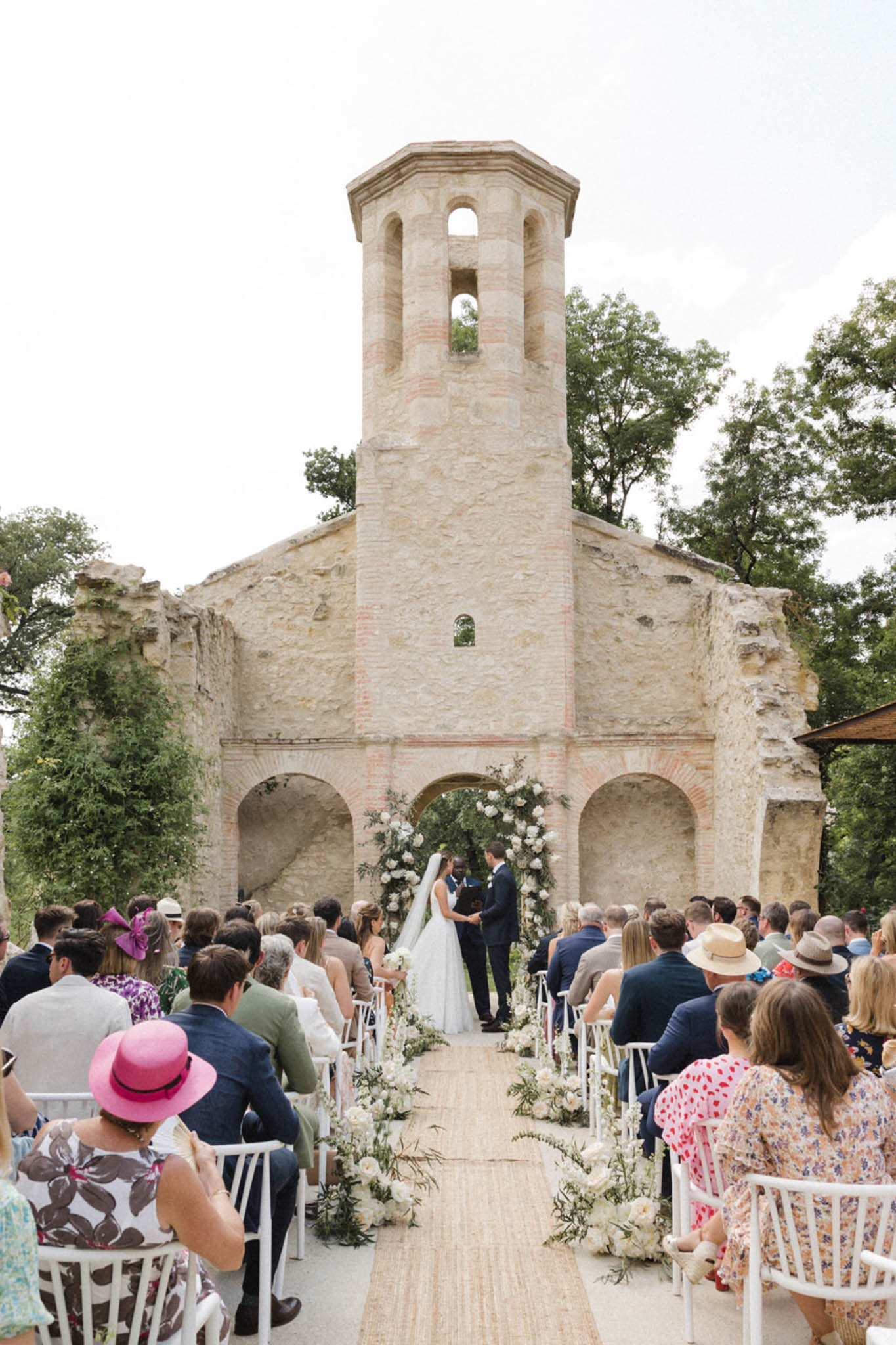 An outdoor wedding ceremony taking place in front of a roofless stone chapel ruin with a distinctive octagonal bell tower and Romanesque arched arcades. The couple stands at the altar beneath a large floral arch composed of white roses, white peonies, and trailing greenery, framed by the central arch of the ruin. A natural jute or woven sisal aisle runner leads from the foreground to the altar, flanked on both sides by clusters of white floral arrangements with trailing foliage placed at the base of white wooden chairs. Approximately 80–100 guests are seated in rows on either side of the aisle, dressed in colorful summer attire including floral prints, a bright pink wide-brim hat, and navy suits. The bride wears a white gown with a long veil, and the officiant and groom stand facing her. The overall floral palette is white and ivory with soft green foliage, and the styling is classic with a relaxed, garden-party feel. Wide shot taken from the back of the aisle looking toward the altar and ruin facade. Potential venue feature image.