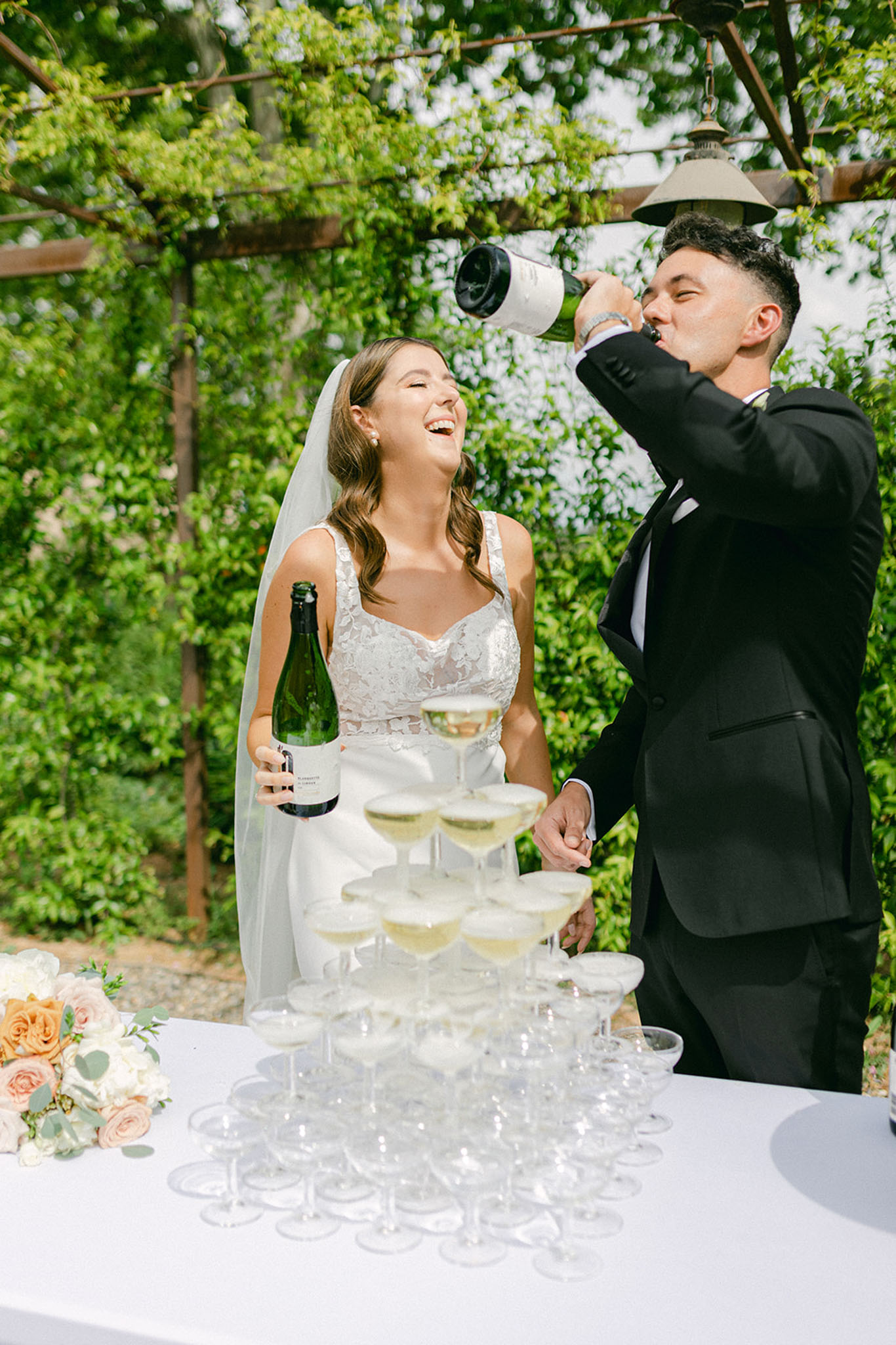 Bride and groom laughing behind a champagne tower under a vine-covered pergola, groom drinking from a bottle