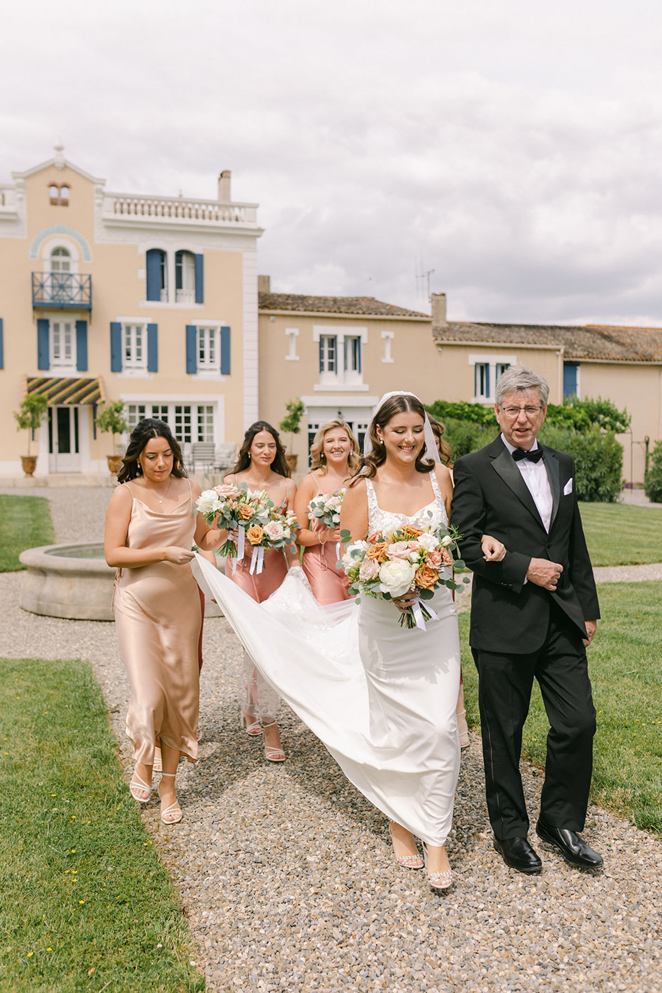 Bride with cathedral veil escorted by father followed by bridesmaids in champagne and dusty rose before manor