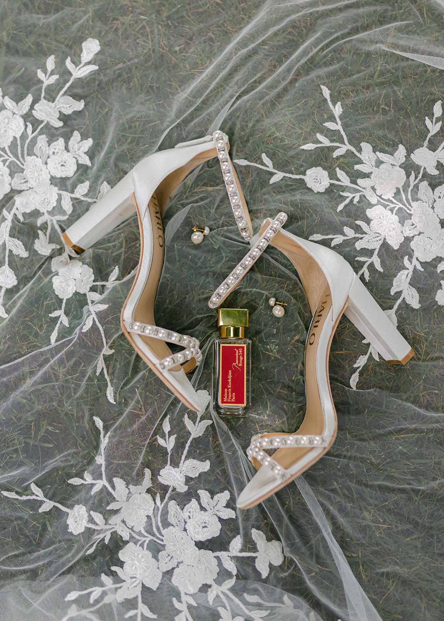 Pearl-strap block heels, Baccarat Rouge 540 perfume, and gold earrings on embroidered tulle veil on grass