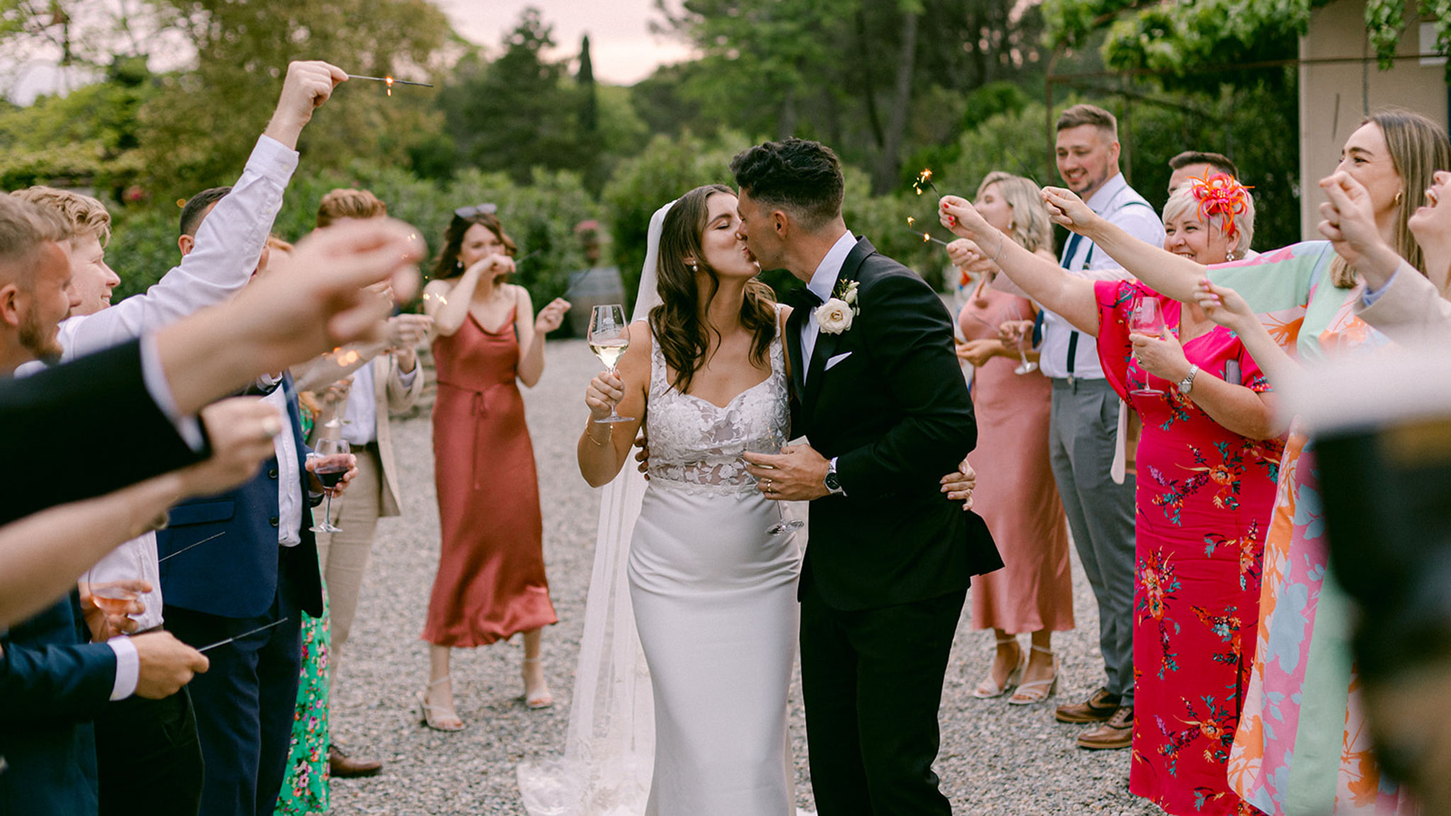 Couple kissing through sparkler send-off tunnel with colourfully dressed guests on gravel path