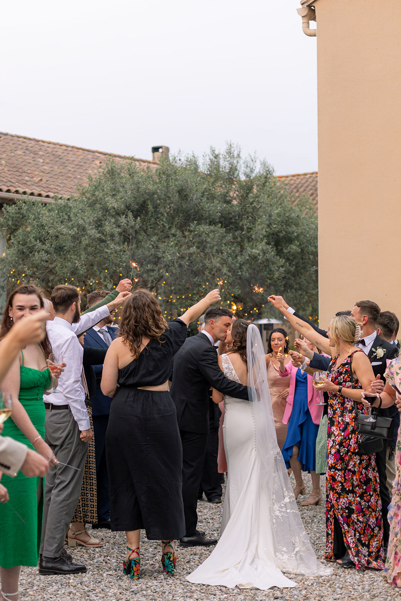 Sparkler send-off with bride and groom kissing as guests form a lit archway on a gravel courtyard beside a French farmhouse