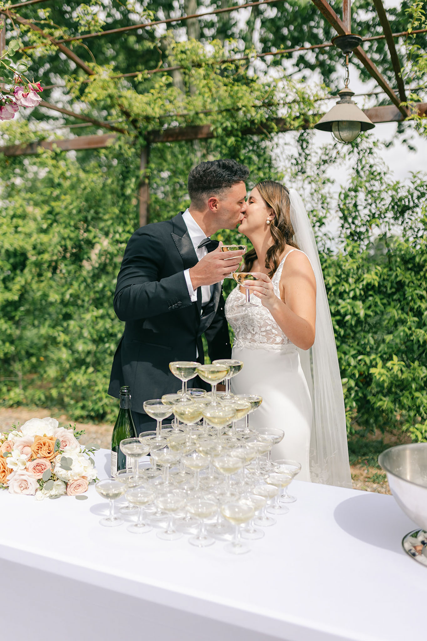 Couple kissing at champagne tower under vine-covered pergola with blush and peach rose arrangement