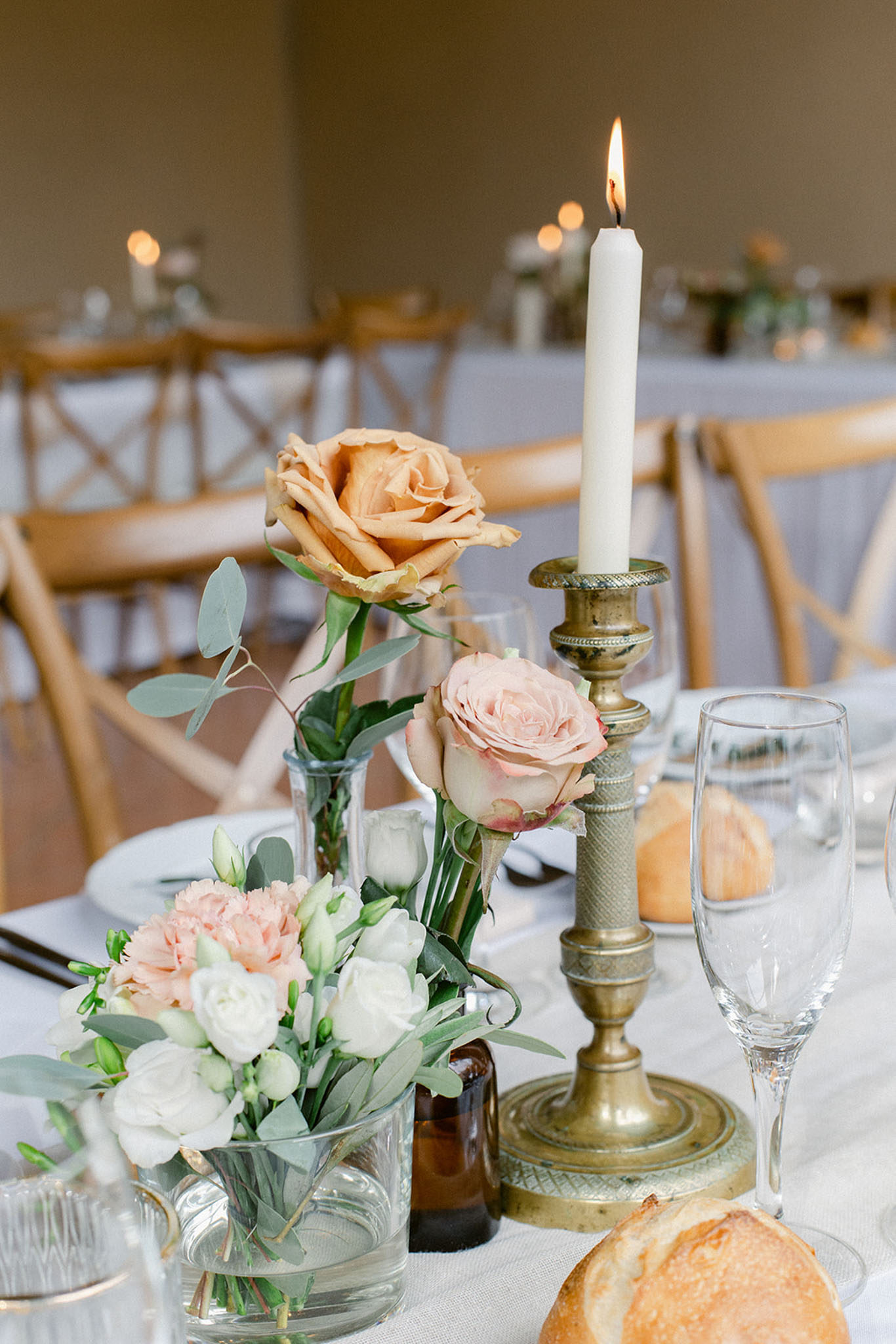 Close-up detail shot of an indoor wedding reception table centerpiece. The arrangement features a low glass bowl with blush pink carnations, white lisianthus buds, and eucalyptus foliage, alongside a clear glass bud vase holding a peach-toned rose with eucalyptus stems and a blush pink garden rose. A brass vintage-style candlestick holds a lit white taper candle. Small amber glass bottles are also incorporated into the styling. The table is set with white linen, crystal champagne flutes, wine glasses, and bread rolls are visible. Additional tables set with matching decor and lit candles are softly blurred in the background, with natural wood cross-back chairs throughout. The overall decor palette is warm and muted — peach, blush, ivory, and brass — with a classic yet relaxed styling approach.