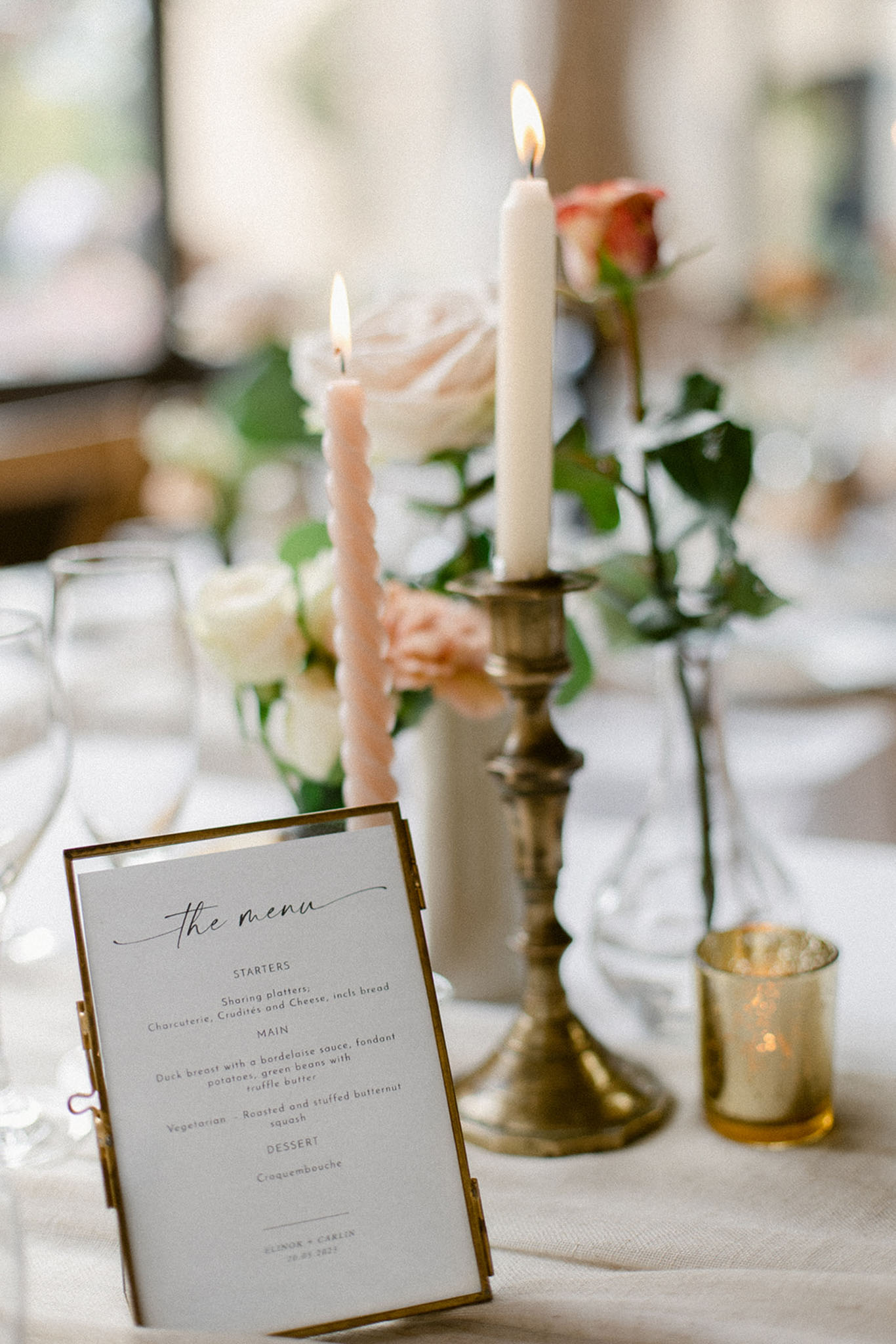 Close-up detail shot of a wedding reception table setting. In the foreground, a printed menu card titled 'the menu' is displayed in a small gold frame, listing starters of sharing platters with charcuterie, crudités and cheese, a main of duck breast with bordelaise sauce or roasted stuffed butternut squash, and a dessert of croquembouche, personalised with the couple's names at the bottom. Behind the menu, a brass candlestick holds a lit ivory taper candle, alongside a lit blush pink twisted taper candle. The centerpiece includes cream and peach garden roses with green foliage, a small gold mercury glass votive candle holder, and clear glassware on a white linen tablecloth. The overall decor palette is ivory, blush, peach, and antique gold, consistent with a classic, romantic styling approach.