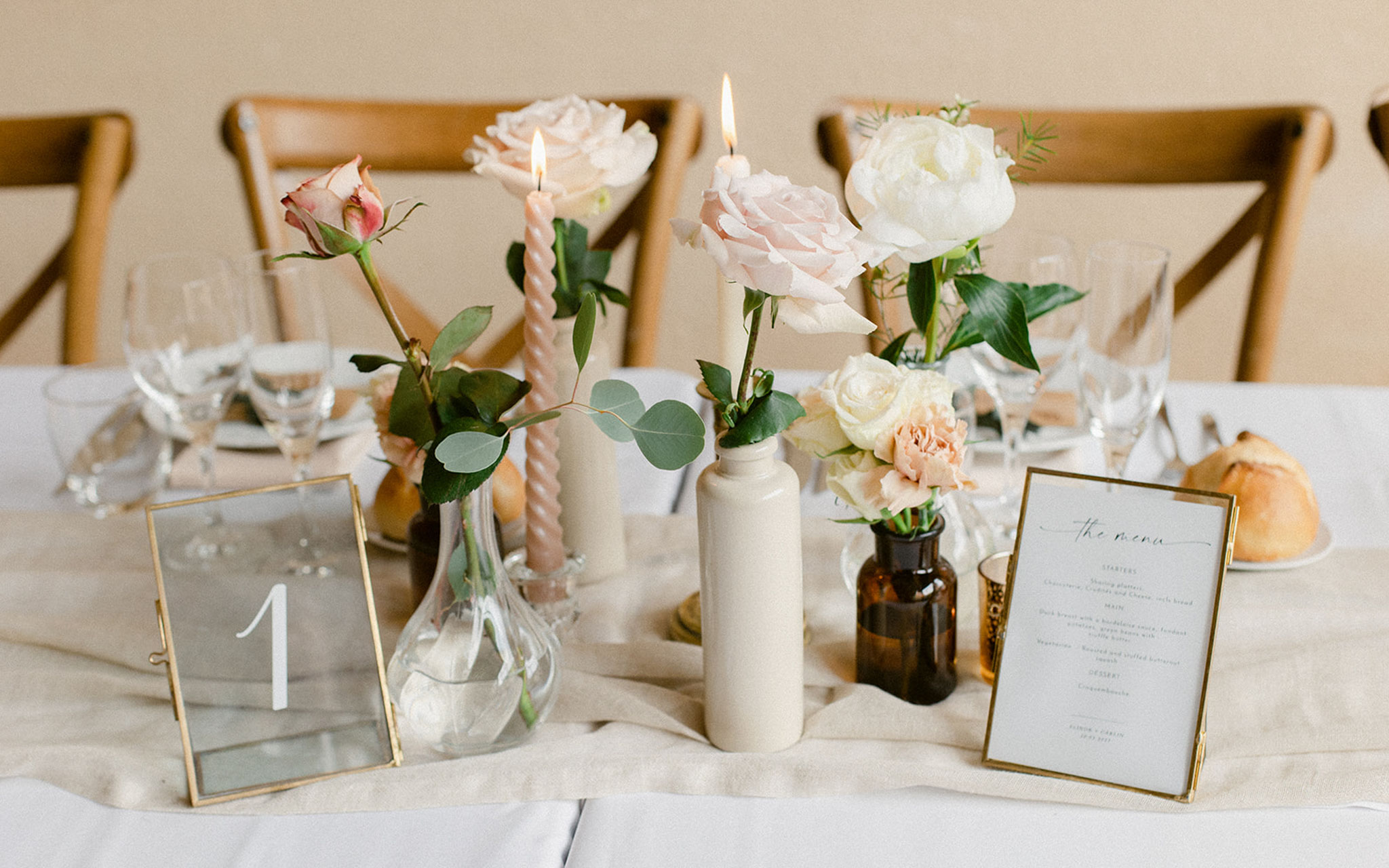 Reception table centerpiece with mismatched bud vases holding blush roses, twisted taper candles, and gold-framed table nu...
