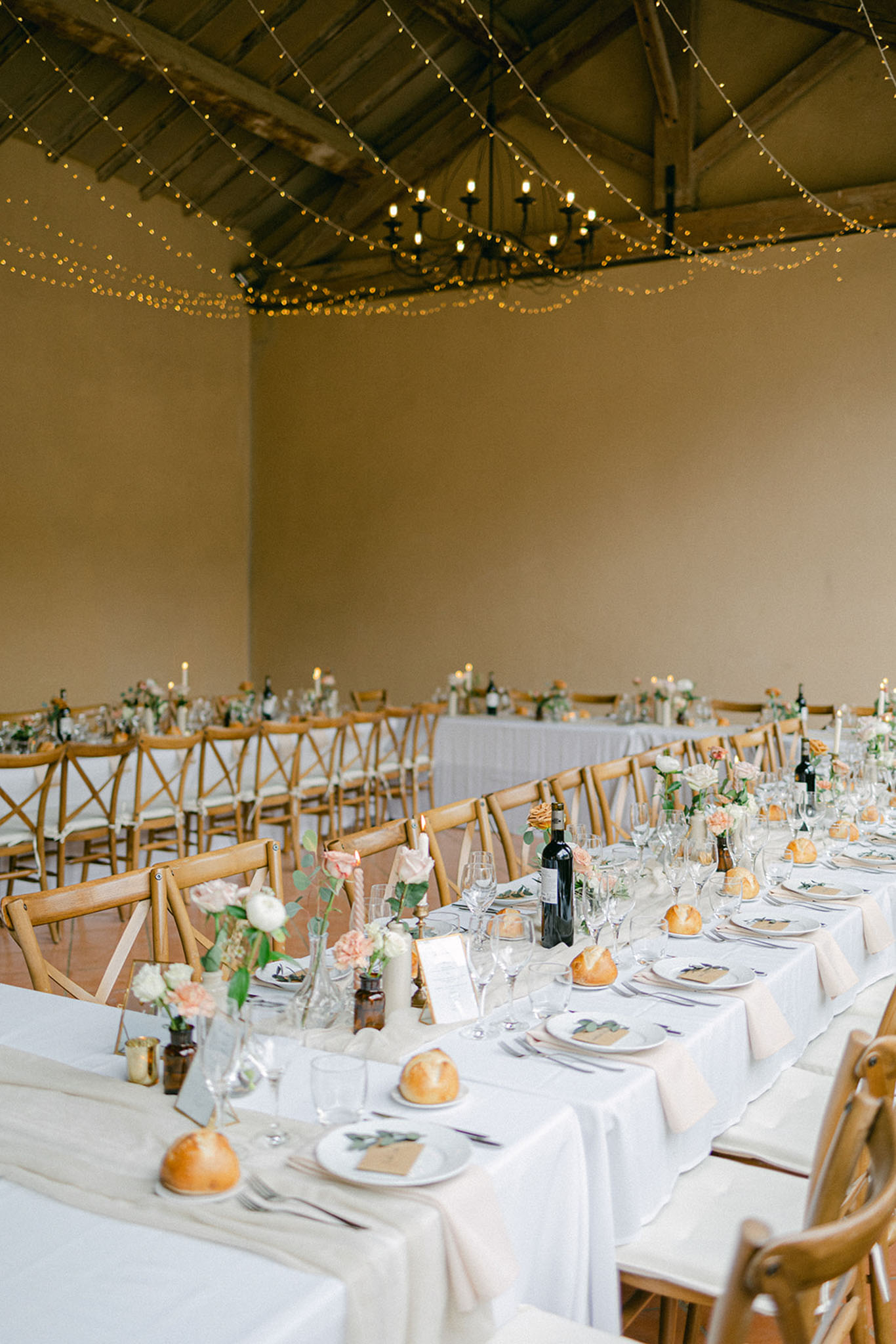 Rustic barn reception with long tables, blush roses in bud vases, twisted candles, and fairy-lit wooden beams