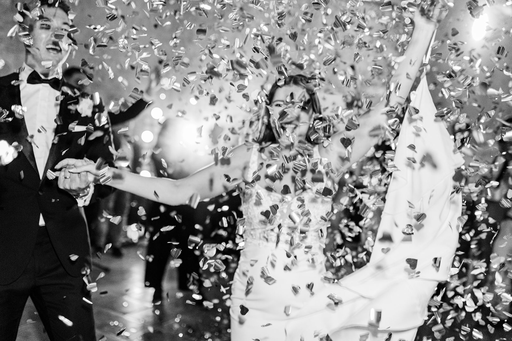 Black and white photo of bride and groom laughing on the dance floor surrounded by heart-shaped confetti