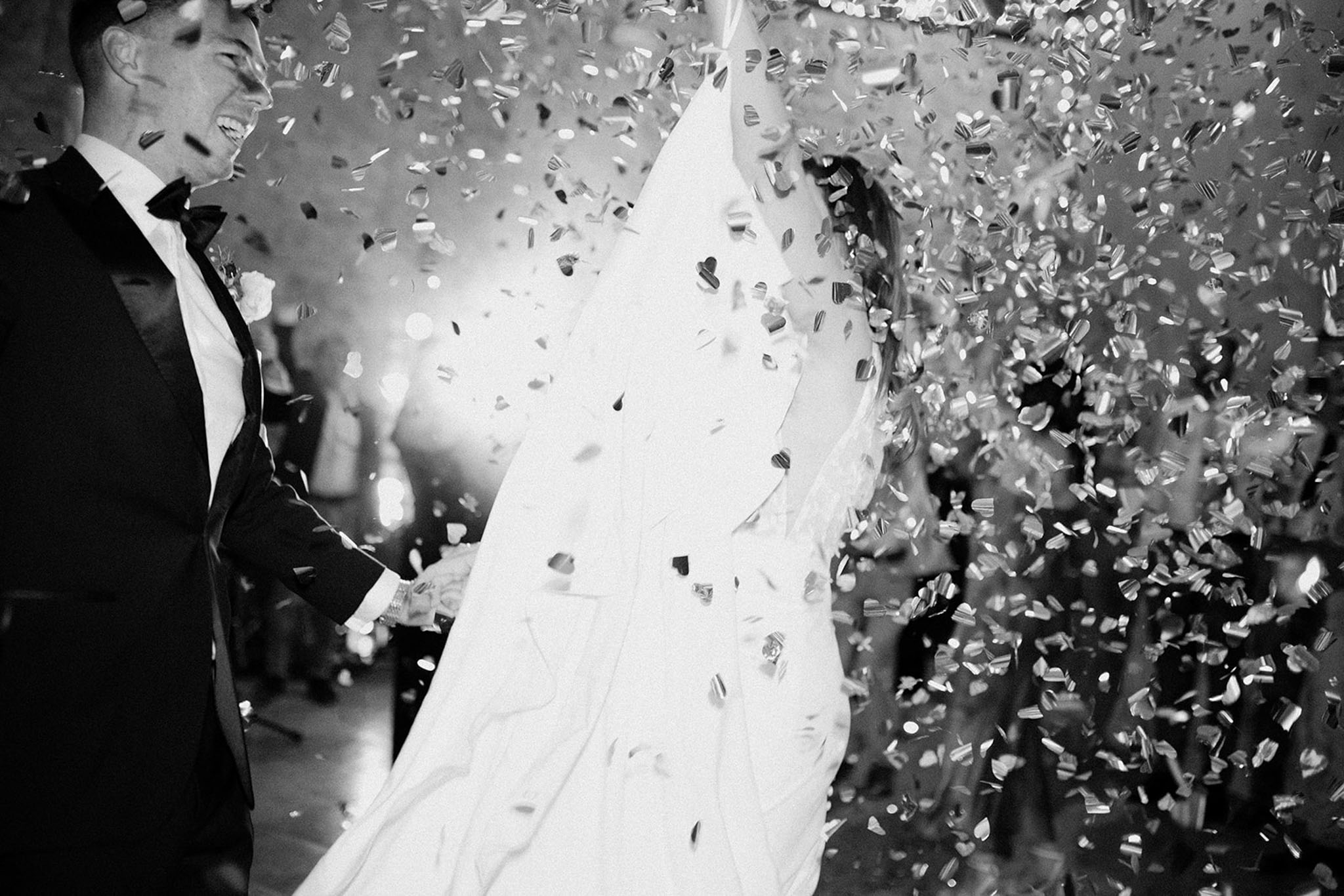 This is a black-and-white image capturing a couple during a reception dance floor moment, surrounded by a heavy shower of confetti. The groom, positioned on the left, wears a dark tuxedo with a bow tie and is laughing with his eyes closed, while the bride spins in a full-skirted white gown, her dress caught mid-motion with movement blur. The confetti pieces — appearing as a mix of small shapes including what look like heart and rectangular cut pieces — fill nearly the entire frame in high contrast against the dark background. The image is shot at close range, cropping both figures partially, with the motion blur and confetti density creating a high-energy, celebratory composition.