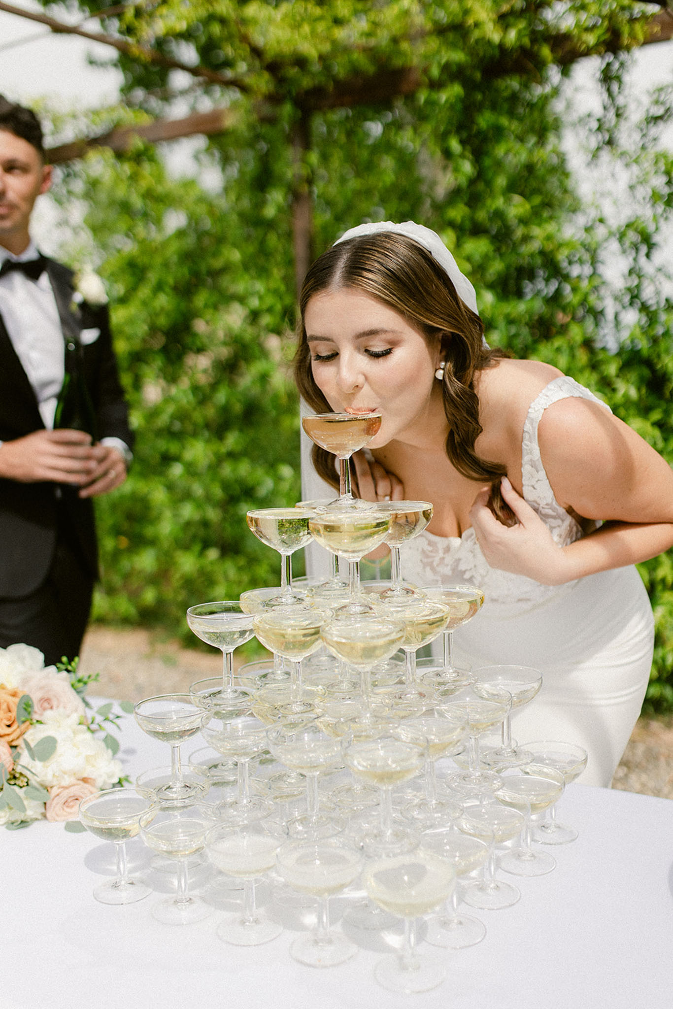 Bride sipping from the top glass of a champagne tower outdoors under a vine-covered pergola