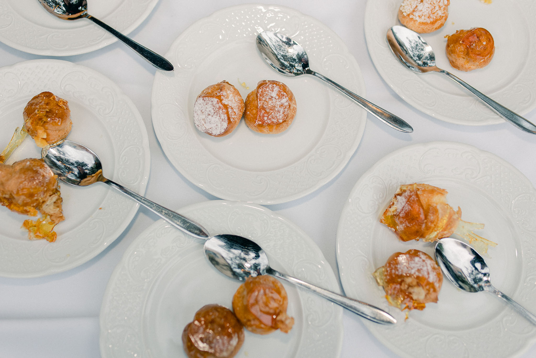 Close-up overhead shot of multiple white embossed dessert plates arranged on a white linen tablecloth, each holding two profiteroles or cream puffs dusted with powdered sugar and glazed with caramel, accompanied by silver dessert spoons. Several plates show the pastries partially eaten, indicating the dessert course is underway during the wedding reception. The plating is consistent across all visible plates, suggesting a formal seated dinner service. The white-on-white palette of the crockery and linen creates a clean, minimal backdrop for the golden-brown pastries.