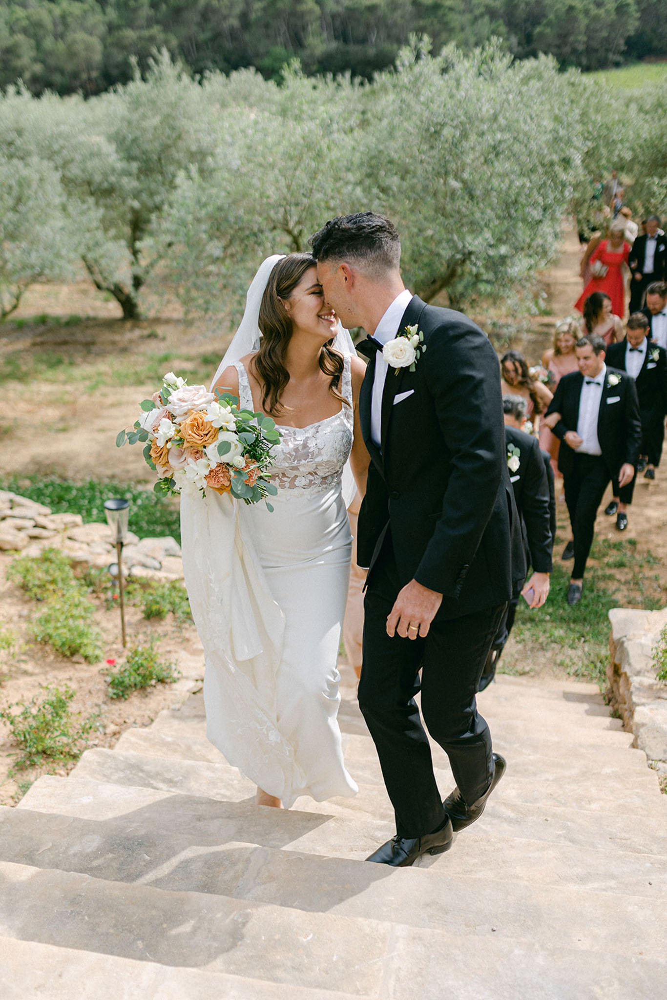 Couple touching foreheads on olive grove steps bride with amber and blush bouquet groomsmen and guests behind