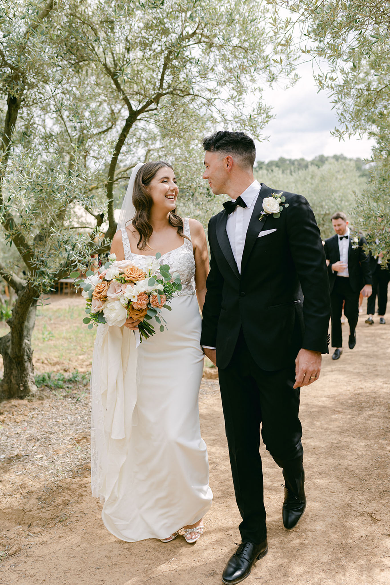 Bride and groom walking hand in hand through olive grove, bride holding terracotta and white bouquet with cathedral veil