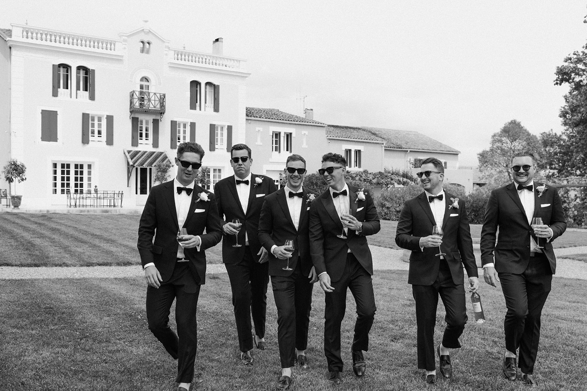 Six groomsmen in dark tuxedos walk across chateau lawn laughing in black and white photo