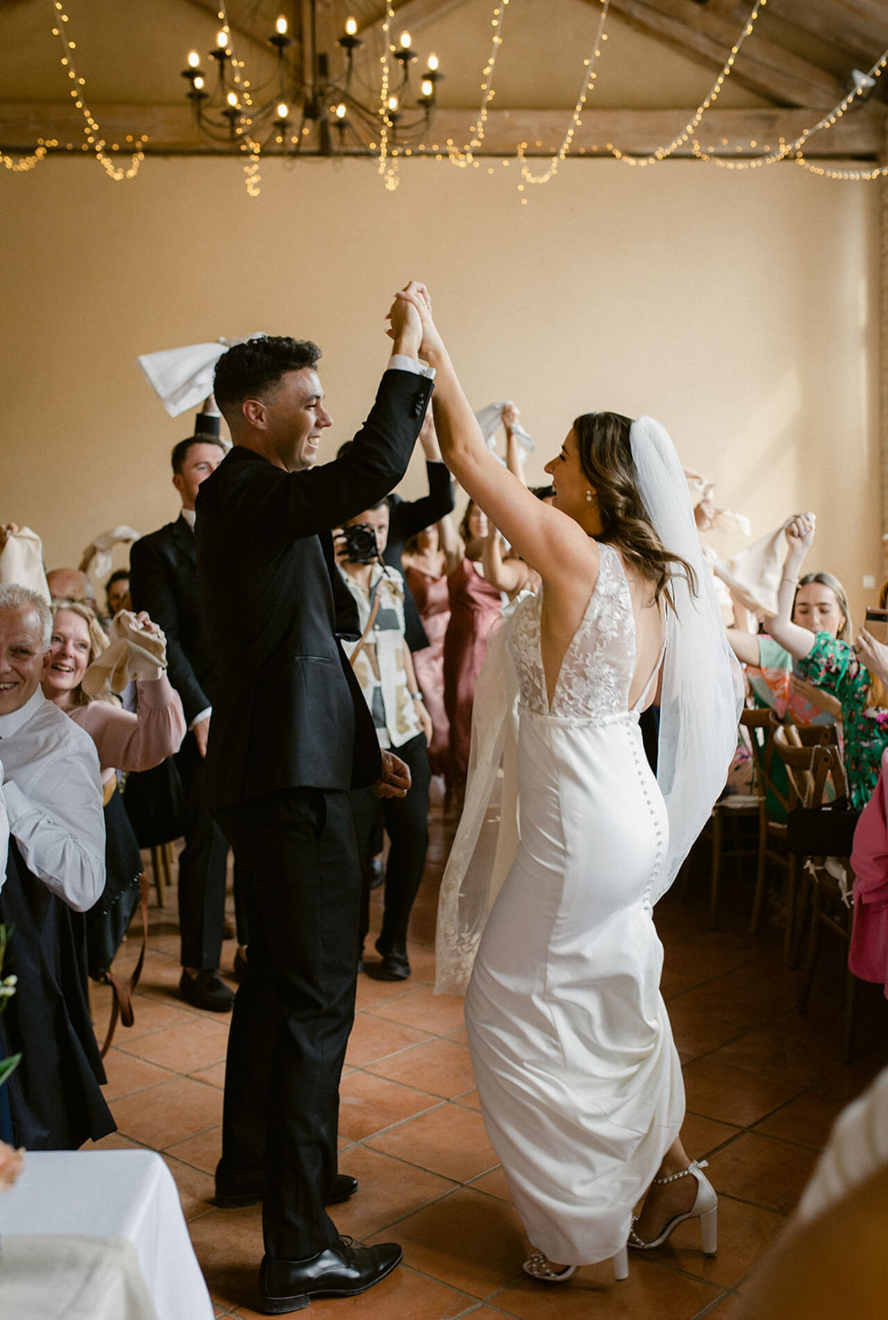 The bride and groom are dancing and celebrating during their reception entrance inside a warm-toned indoor venue with terracotta tile flooring, exposed wooden ceiling beams, a wrought-iron chandelier, and fairy lights strung across the ceiling. The groom wears a black suit with black dress shoes, and the bride wears a white fitted gown with a lace bodice, open back with button detail down the spine, a cathedral-length veil, and white block-heel sandals. Guests surrounding them — approximately 20–25 visible — are waving white napkins in the air in celebration, a tradition common at French and Mediterranean receptions. The shot is a medium wide portrait-style image taken from a slight low angle, capturing the energy of the room with a photographer visible in the background.