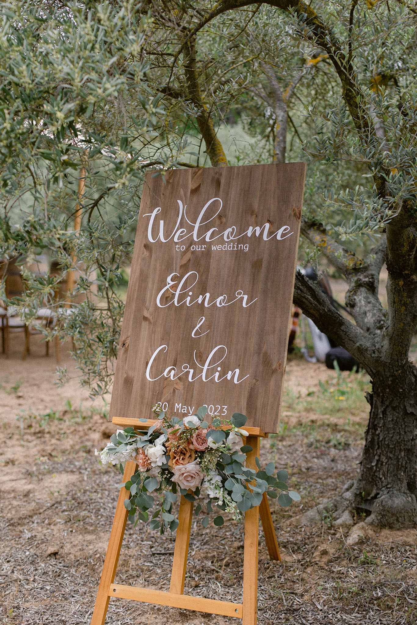 A close-up detail shot of a wooden welcome sign displayed on a natural wood easel at an outdoor wedding ceremony. The sign reads 'Welcome to our wedding, Elinor & Carlin, 20 May 2023' in white hand-lettered calligraphy script on a warm brown wood-grain board. The base of the easel is decorated with a floral arrangement of dusty rose and terracotta roses, ivory blooms, and eucalyptus greenery. The sign is positioned among olive trees, with ceremony seating faintly visible in the background, contributing to a rustic, garden-style aesthetic.