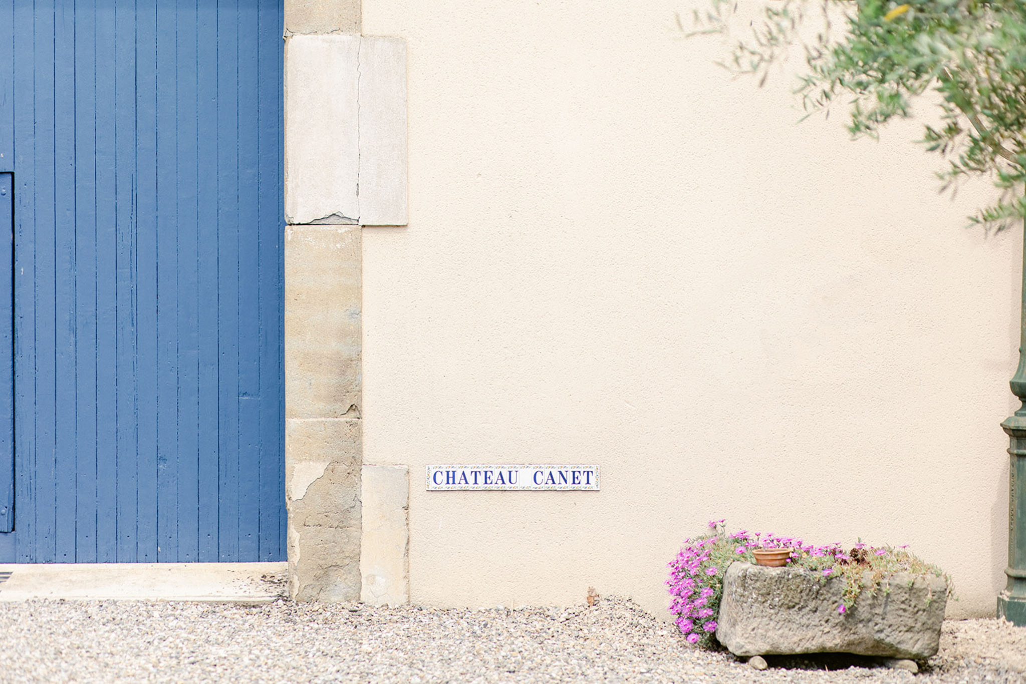 A detail shot of an exterior wall at Château Canet, featuring a blue-and-white ceramic name plaque reading 'CHATEAU CANET' mounted on a cream-rendered wall beside a cobalt blue painted wooden door with stone quoining. A rustic stone trough planter at the base of the wall holds bright magenta-pink small flowers, and a tree with green foliage is partially visible in the upper right corner. The foreground shows a gravel surface. Potential venue feature image.