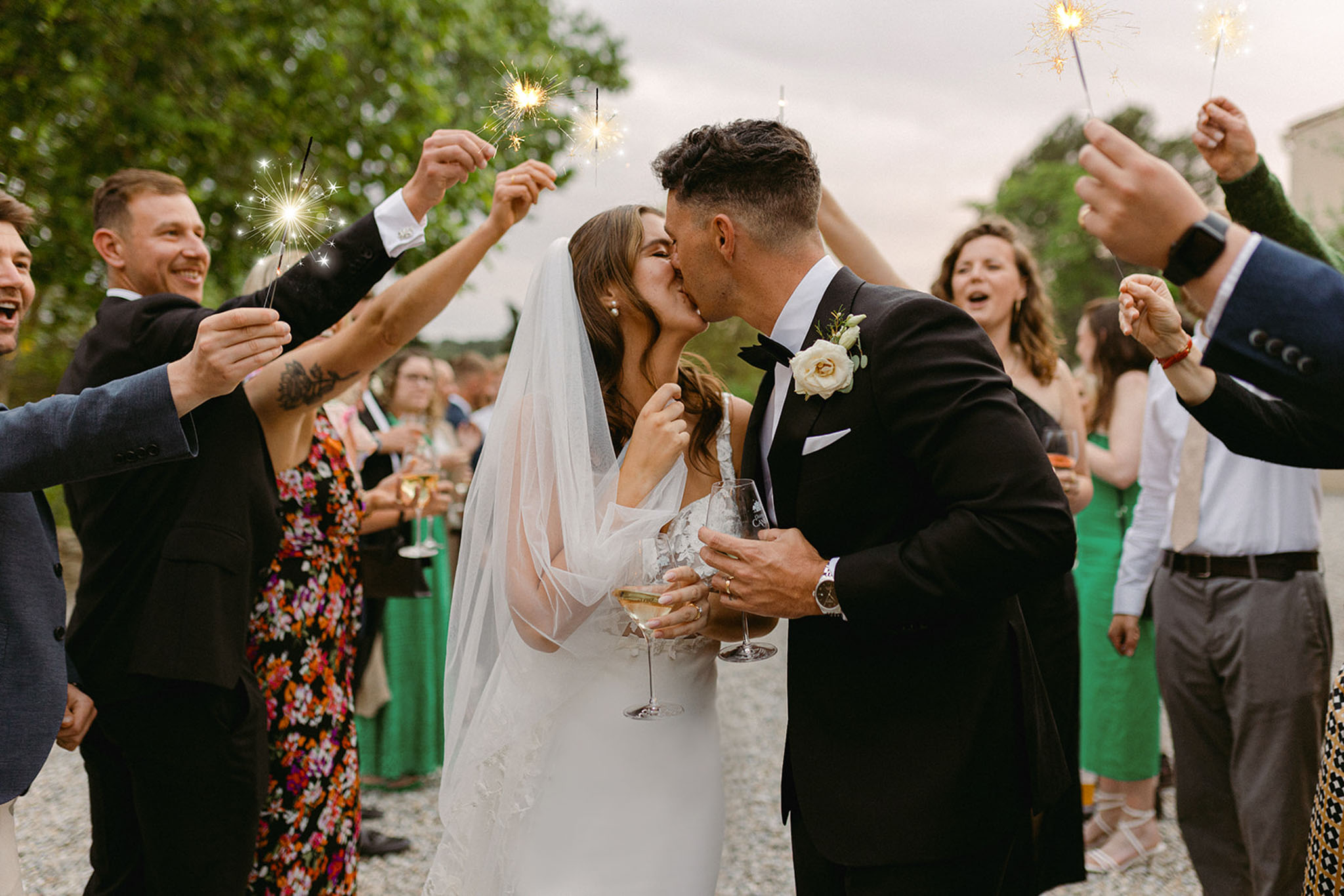 Bride and groom kiss during outdoor sparkler send-off surrounded by guests holding lit sparklers