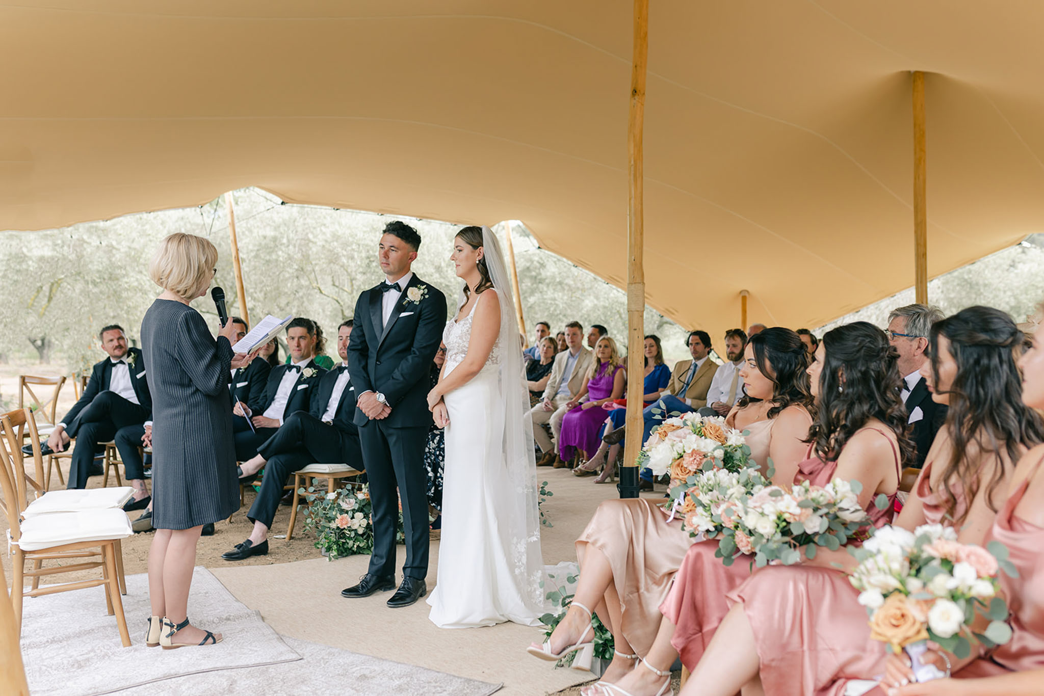 Wedding ceremony under stretch tent in olive grove with bridesmaids in dusty rose dresses holding peach rose bouquets
