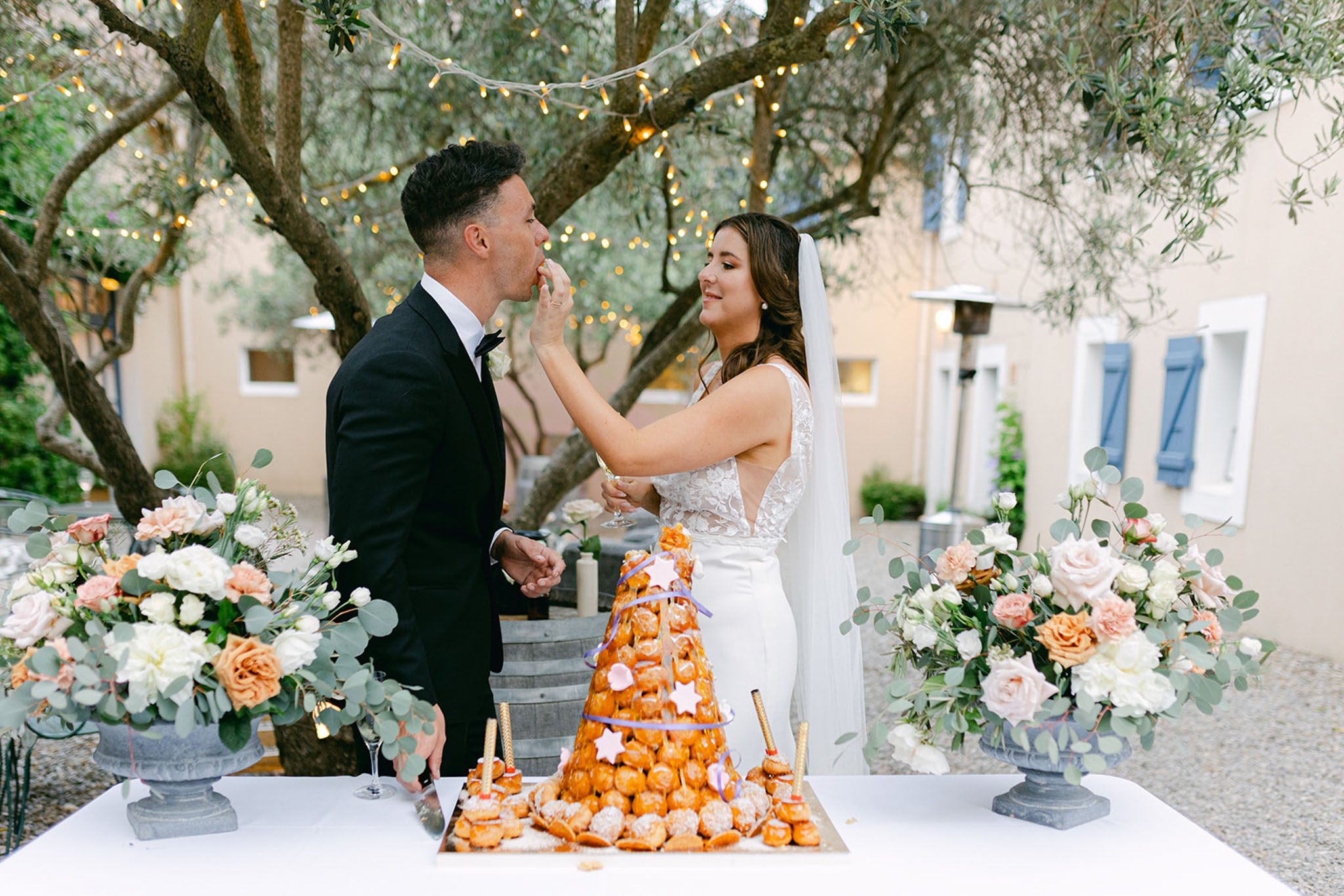 The bride and groom are cutting and sharing their croquembouche during an outdoor reception at a Provençal-style venue with cream-rendered walls and blue shutters visible in the background. The bride wears a sleeveless white lace-bodice jumpsuit with a long veil, while the groom wears a black tuxedo with a bow tie; she is feeding him a profiterole from the tower. The croquembouche is decorated with gold star ornaments, purple ribbon spirals, and gold taper candles, displayed on a white-clothed table flanked by two stone urn arrangements filled with blush and peach garden roses, ivory blooms, and eucalyptus. Warm fairy lights are strung through the tree branches overhead, and the overall decor palette is soft peach, blush, and white with a classic French outdoor reception aesthetic. The shot is a medium portrait-style image taken at eye level.