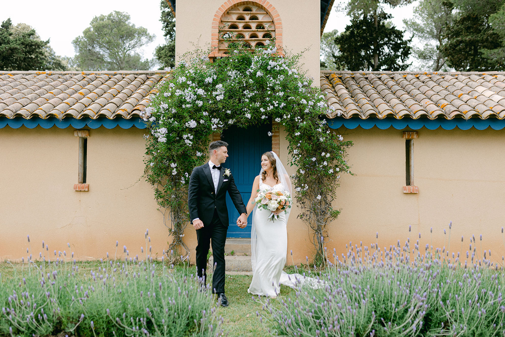 A couple portrait taken outdoors in front of a Provençal-style building with terracotta roof tiles, blue-painted eaves, and a cobalt blue door framed by a climbing jasmine vine with small white flowers. The groom wears a black tuxedo with a bow tie and a white boutonnière, while the bride wears a fitted ivory slip-style gown with a cathedral-length veil and carries a rounded bouquet of ivory roses, peach blooms, and eucalyptus. The couple hold hands and look at each other as they walk, with rows of purple lavender in the foreground adding a distinctly South of France character to the setting. The shot is a medium-wide portrait with the architectural backdrop centered behind them.