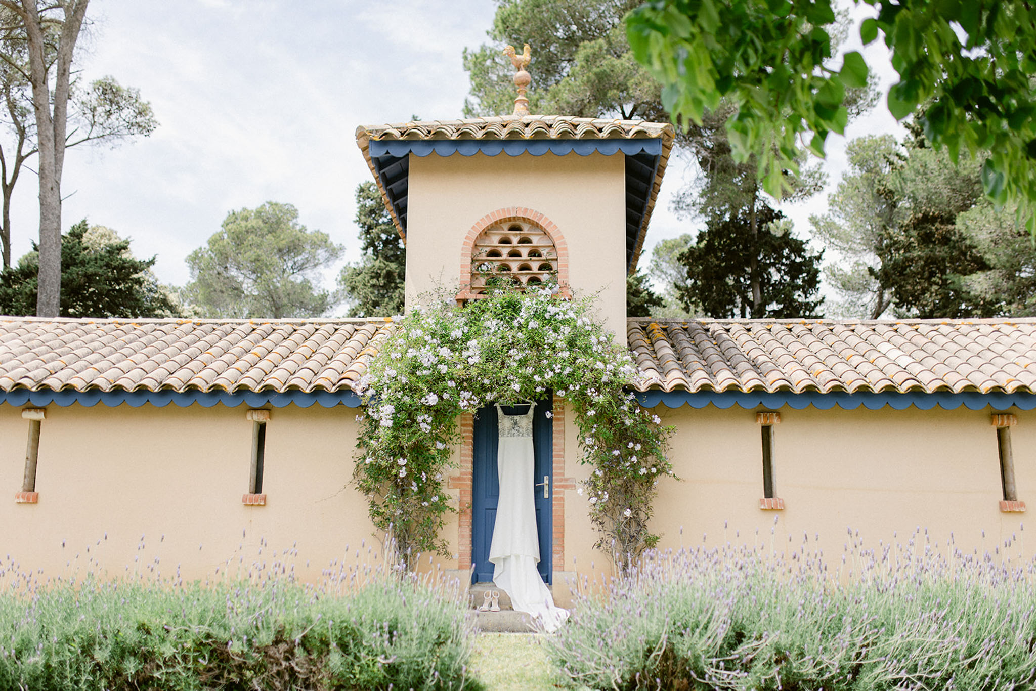 A white wedding dress with what appears to be a fitted bodice and flowing skirt is hung on a deep navy blue door of a Provençal-style outbuilding, used as a getting-ready detail shot. The building features warm sand-colored rendered walls, terracotta roof tiles with navy scalloped trim, and a central tower topped with a gold weathervane rooster. A climbing plant with small pale lavender-white flowers cascades over the doorway and tower. The foreground features rows of purple lavender in bloom. This is a wide establishing shot that incorporates the dress as a styling detail within the architectural setting. Potential venue feature image.