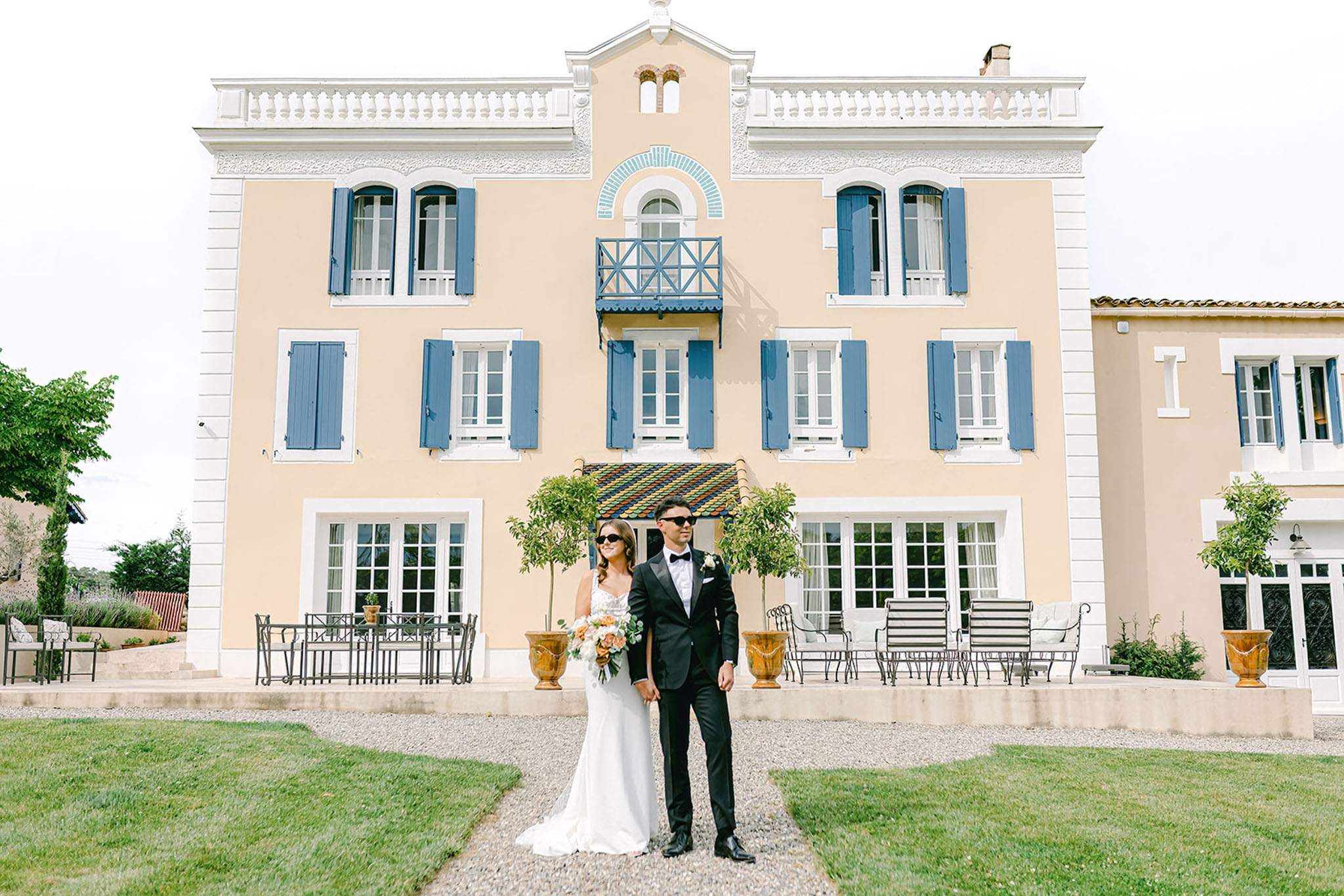 Bride and groom with sunglasses before peach Provencal villa with topiary and glazed tile roof