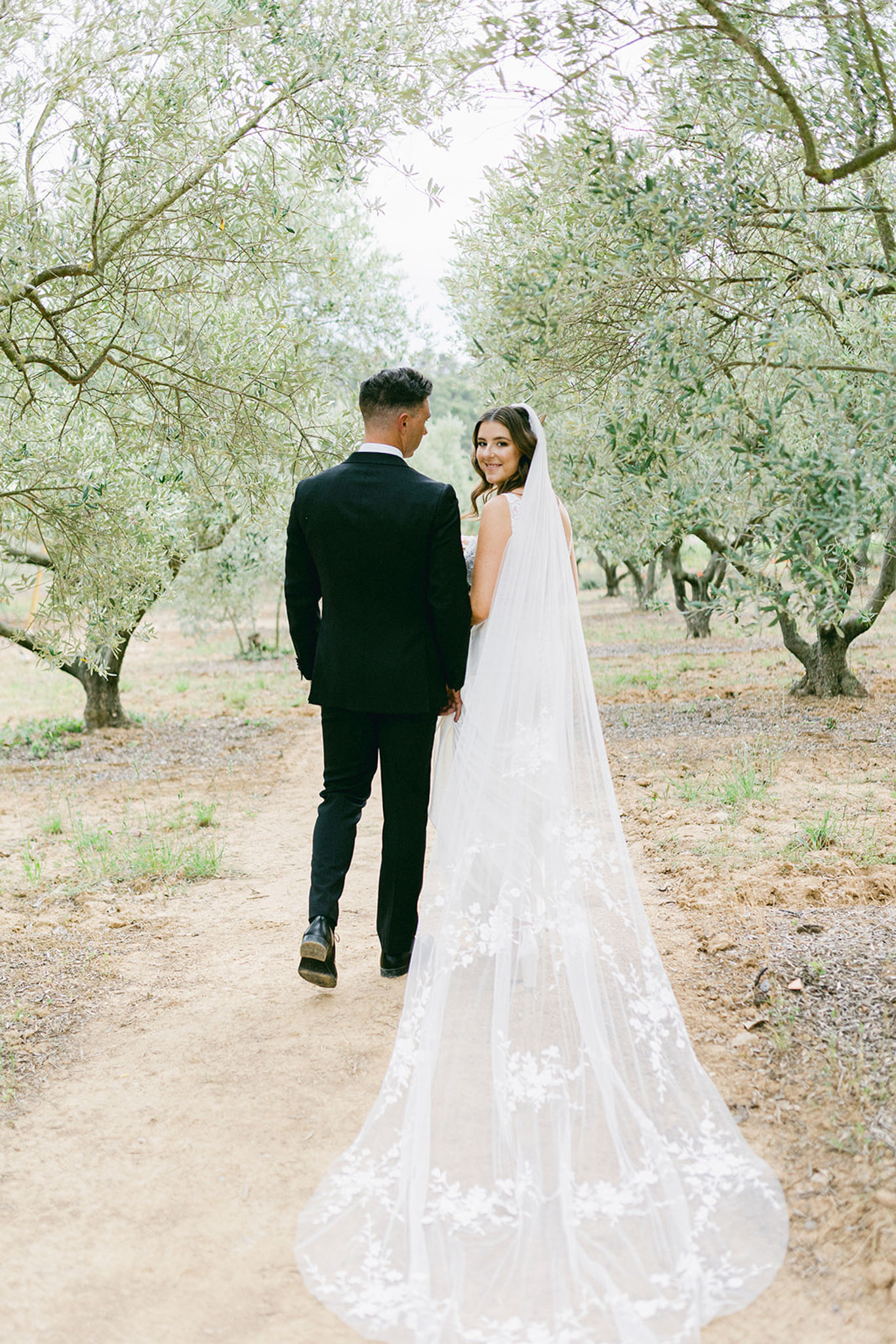 Couple walks away through olive grove as bride turns back smiling with applique-edged cathedral veil trailing