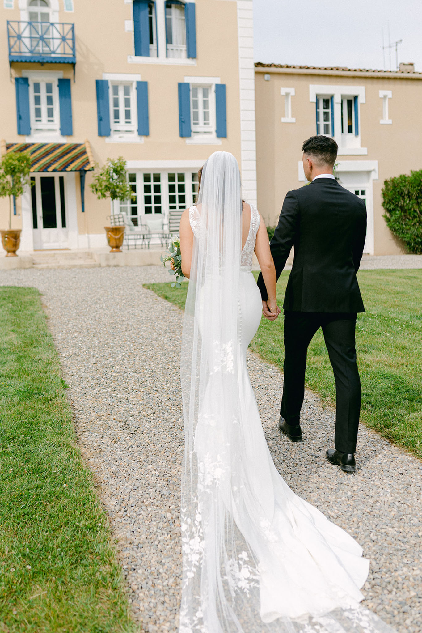 Bride and groom walking hand in hand toward ochre manor house with blue shutters, lace train trailing behind