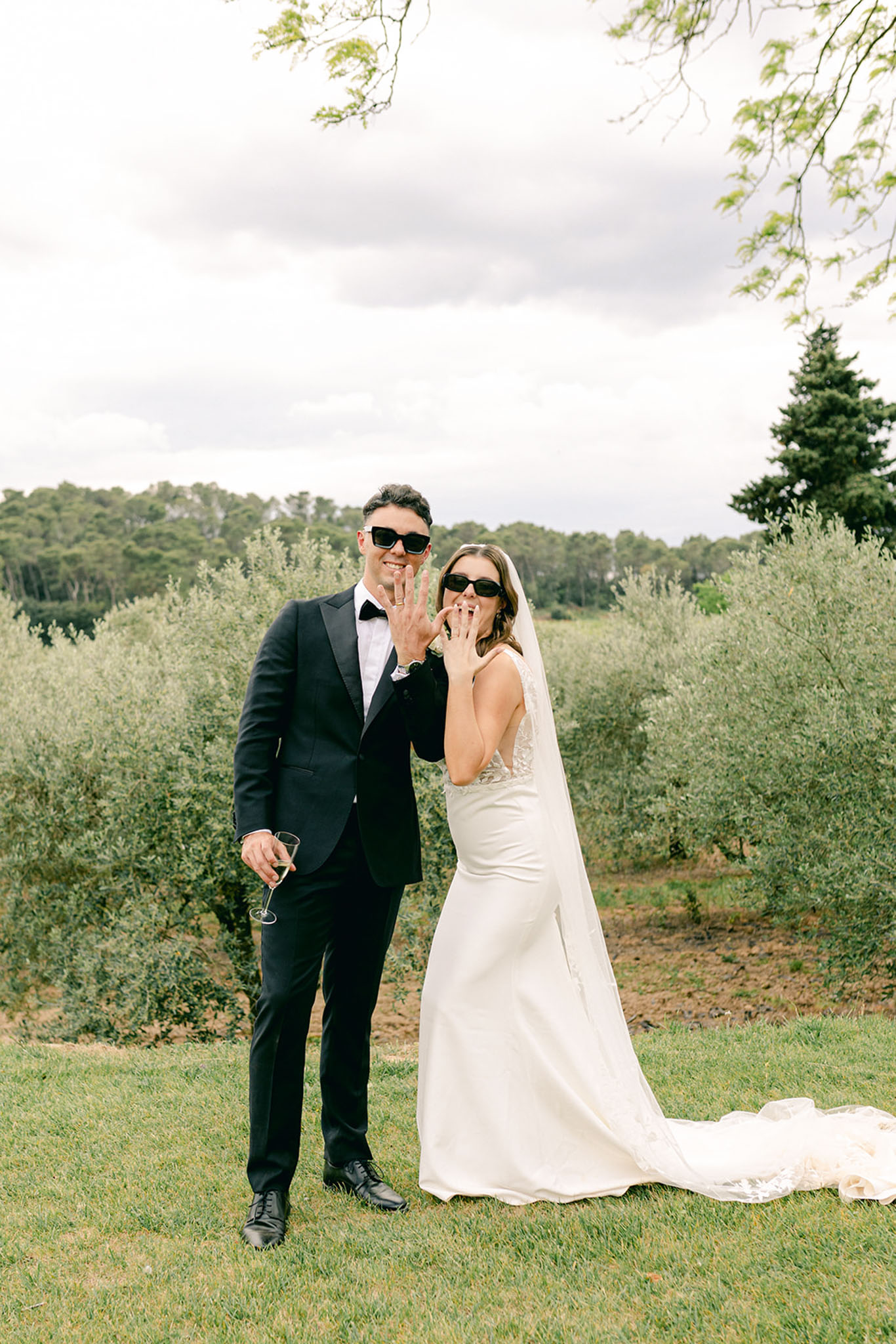 Couple showing wedding rings in matching sunglasses with cathedral lace veil before olive tree row