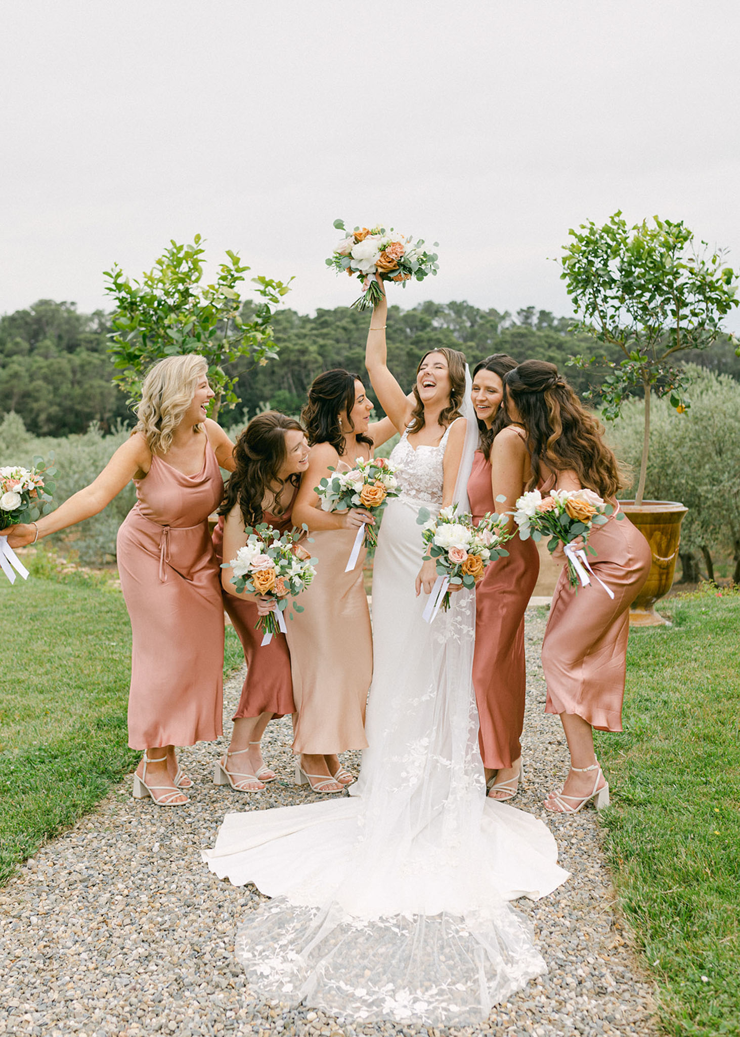 Bride in white lace gown with five bridesmaids in terracotta and champagne satin dresses on a gravel path