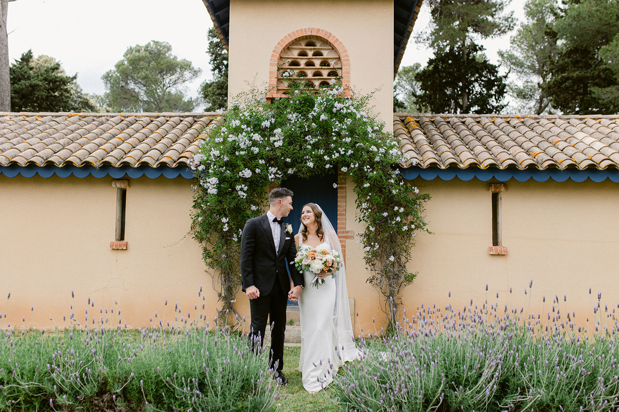 A couple portrait taken outdoors at what appears to be a Provençal-style property, with the bride and groom standing hand-in-hand in front of a ochre-rendered building featuring terracotta roof tiles with blue scalloped trim and a decorative brick arched niche above a blue door. The groom wears a black tuxedo with a bow tie and a white boutonnière, while the bride wears a fitted ivory sleeveless gown with a cathedral-length veil and carries a bouquet of peach garden roses, white peonies, and eucalyptus. A climbing white-flowered vine frames the doorway behind them, and rows of lavender plants run across the foreground. The composition is a wide portrait shot with the couple centered against the architectural backdrop.