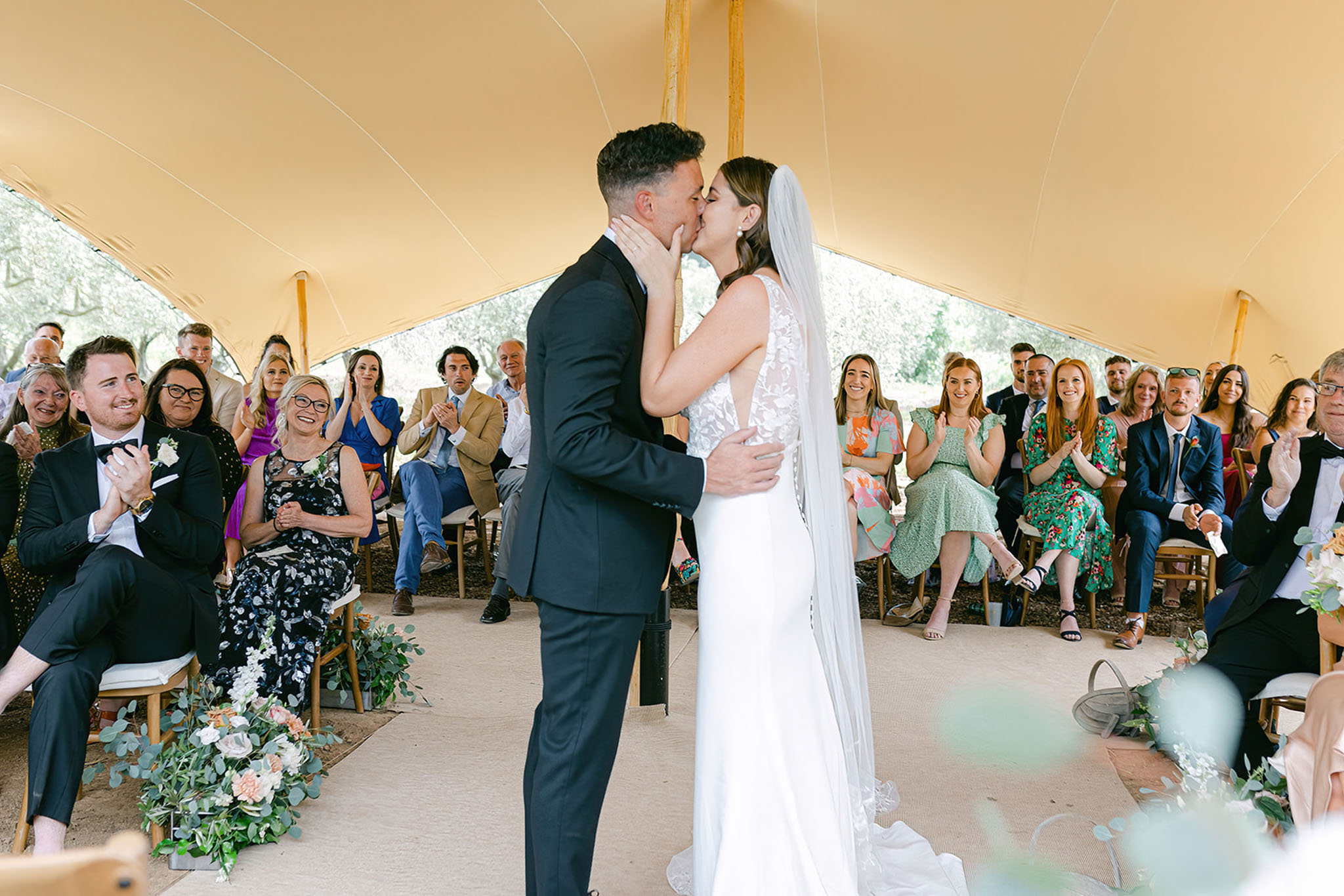 The couple shares their first kiss during an outdoor ceremony held inside an open-sided stretch tent with a warm cream canvas canopy supported by wooden poles. The groom wears a navy suit, and the bride wears a fitted white gown with a lace bodice and a long veil. Approximately 30–40 guests seated on wooden chairs on both sides of the aisle react with applause and smiles. Aisle decor consists of low arrangements of peach and white roses with trailing eucalyptus greenery placed at the base of the seating rows. The wide-angle shot captures the full ceremony space, couple centered in the foreground, with guests filling both sides of the frame.