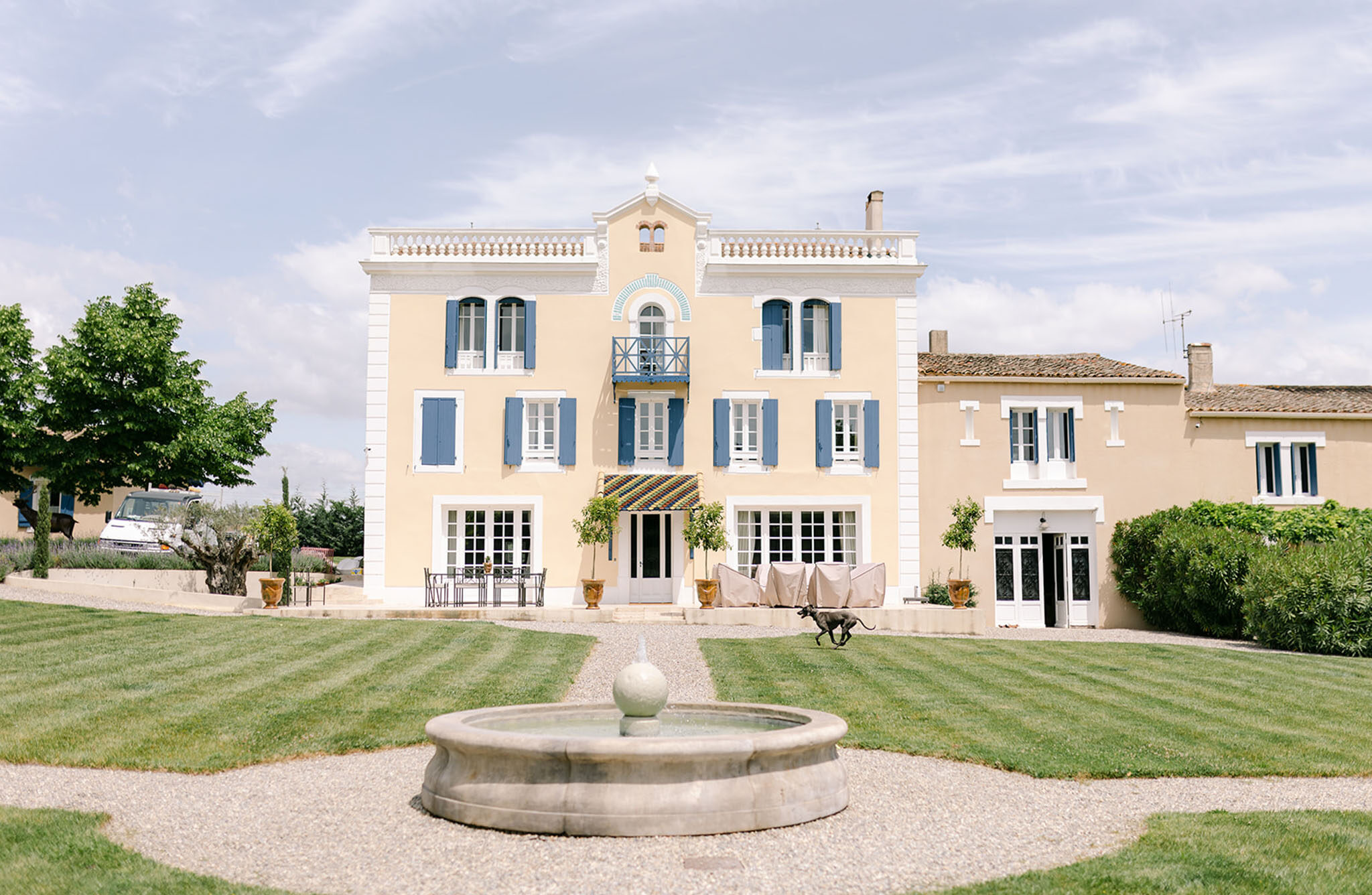 A wide exterior shot of a three-story French manor house with a pale yellow facade, white trim, and blue shutters throughout. The entrance features a colorful geometric-patterned canopy in blue, yellow, and green above the central French doors, flanked by potted topiary trees in terracotta urns. The foreground includes a circular stone fountain with a sphere finial set within a gravel path, surrounded by neatly striped mowed lawn. A dark-colored dog runs across the gravel path in front of the entrance. Outdoor furniture including a metal dining table and chairs is visible to the left of the entrance, with covered furniture to the right. An adjacent lower building in matching yellow render extends to the right. Potential venue feature image.