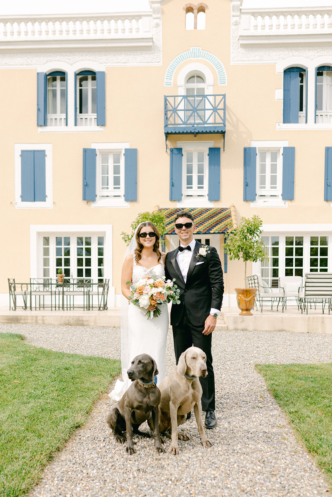 Bride and groom with sunglasses and two dogs before peach villa with blue shutters