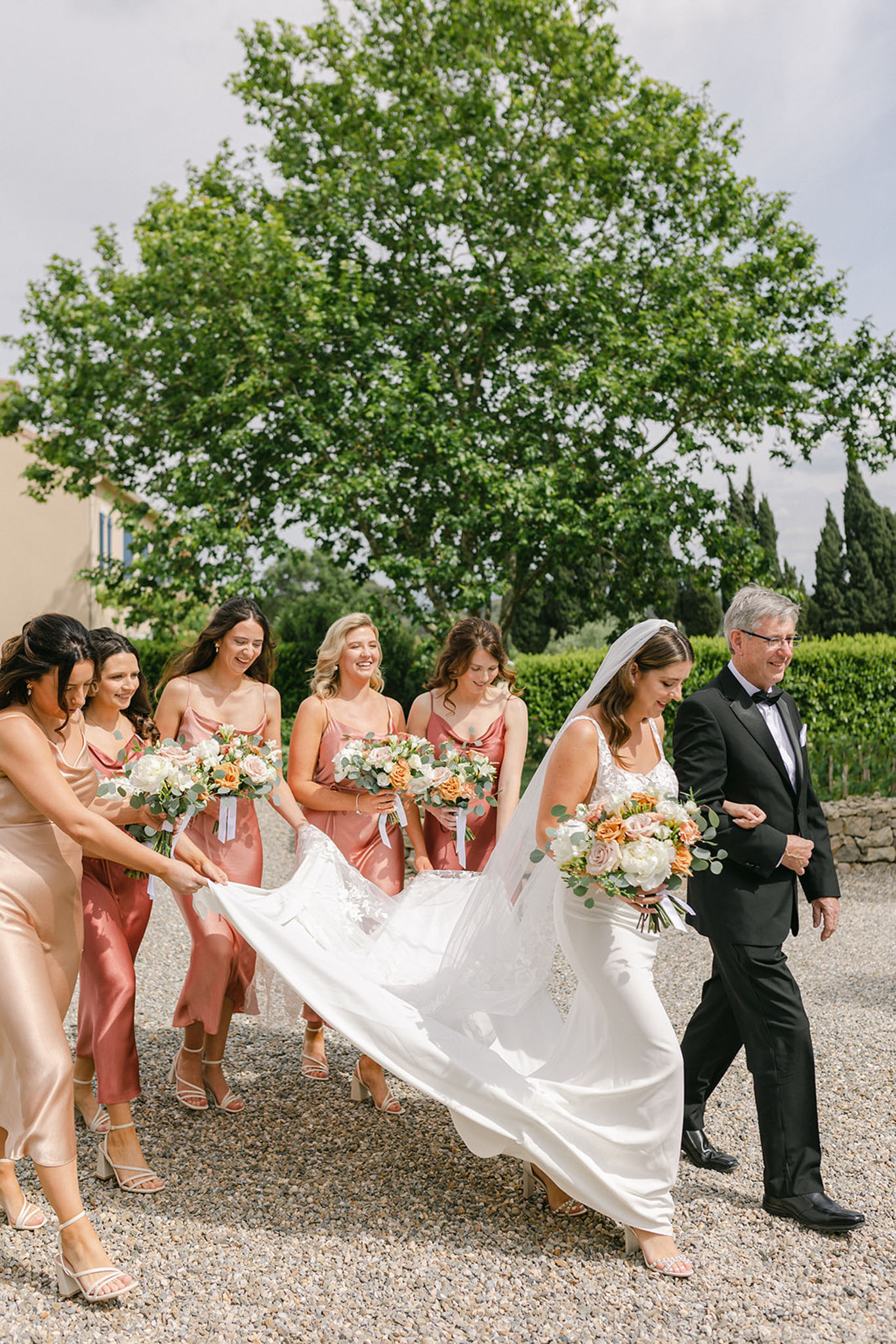 Bride escorted by father with five dusty rose bridesmaids carrying cathedral train on cypress-lined path