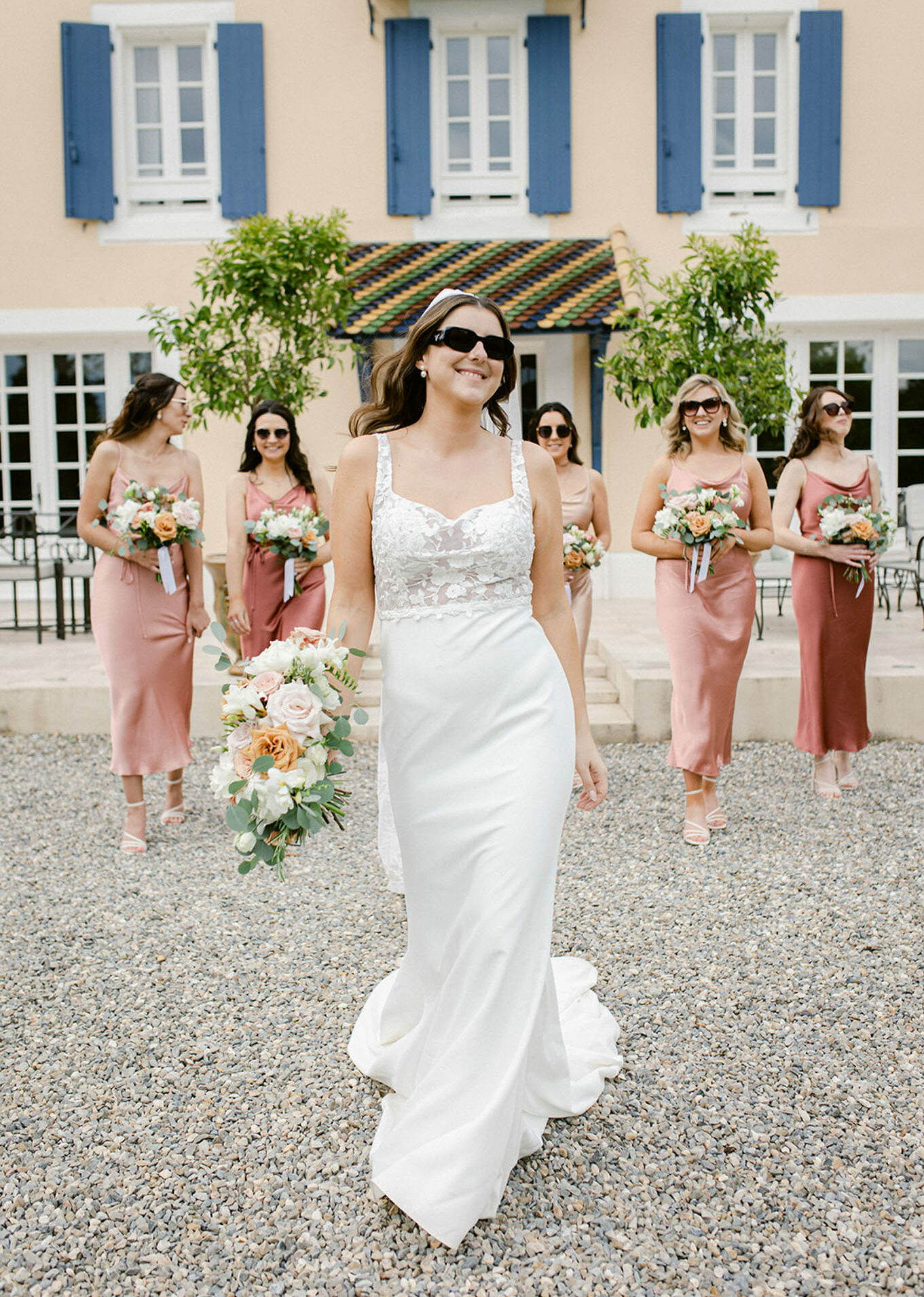 Bride in white lace gown with five bridesmaids in dusty rose satin dresses walking before ochre manor