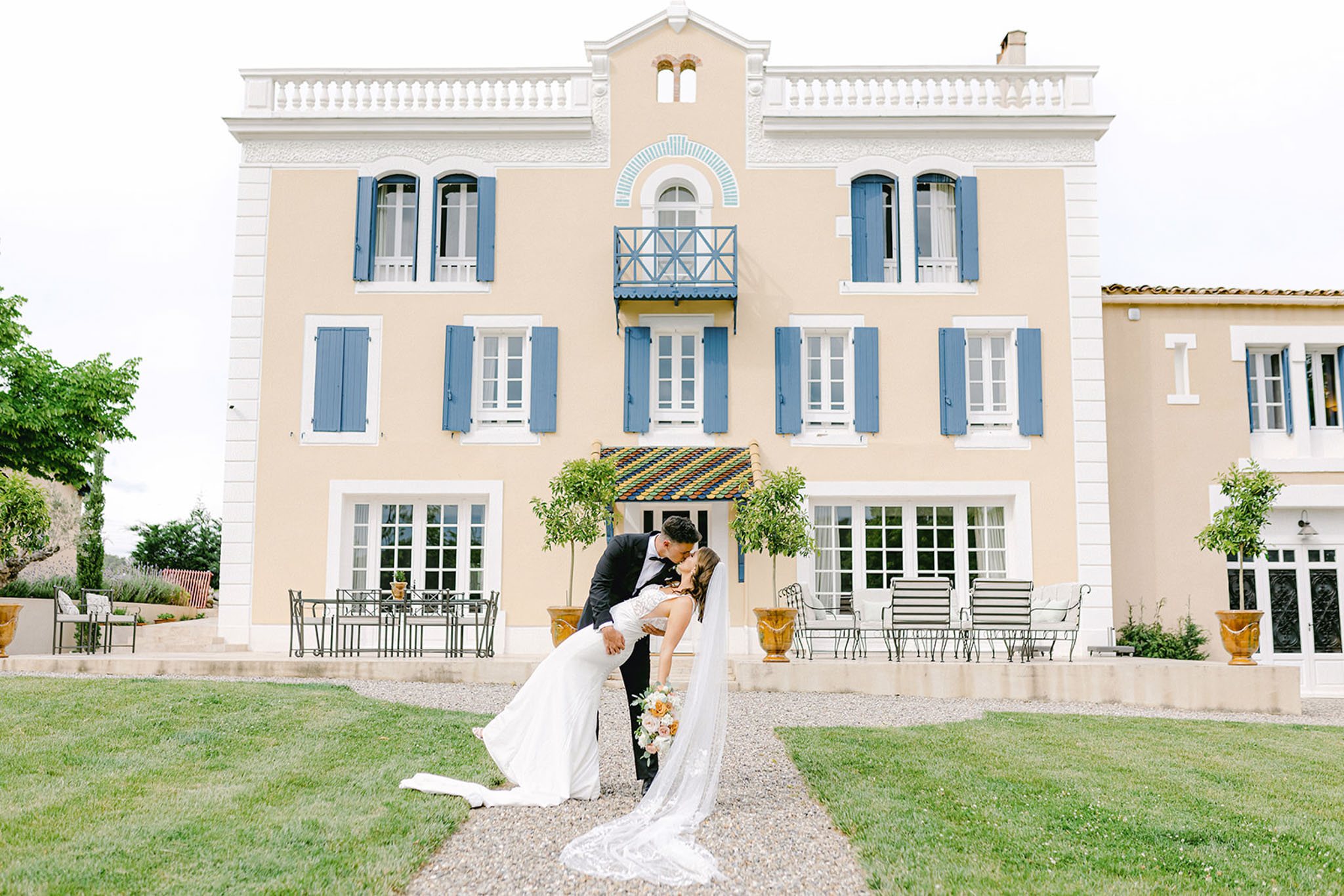 Groom dips bride for a kiss on gravel path in front of peach-facade French villa with blue shutters
