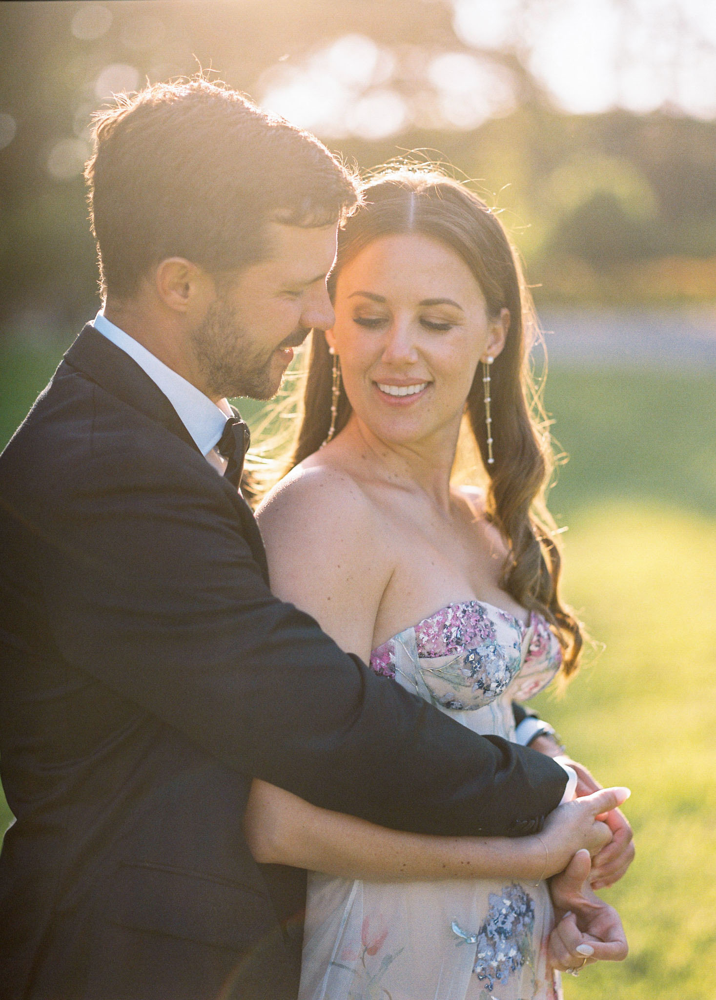 A couple portrait taken outdoors during golden hour, with warm backlit light creating a soft glow around both subjects. The groom, wearing a dark navy suit with a dark tie, stands behind the bride with his arms wrapped around her waist, leaning in close to her. The bride wears a strapless gown with a floral embroidered and sequined bodice featuring pink, blue, and green botanical motifs on a champagne/ivory base, paired with long delicate drop earrings and her dark hair worn in loose waves over one shoulder. Both are smiling and looking downward in a relaxed, candid pose. The composition is a close-up portrait with a shallow depth of field, set against an outdoor lawn setting with a heavily blurred background.