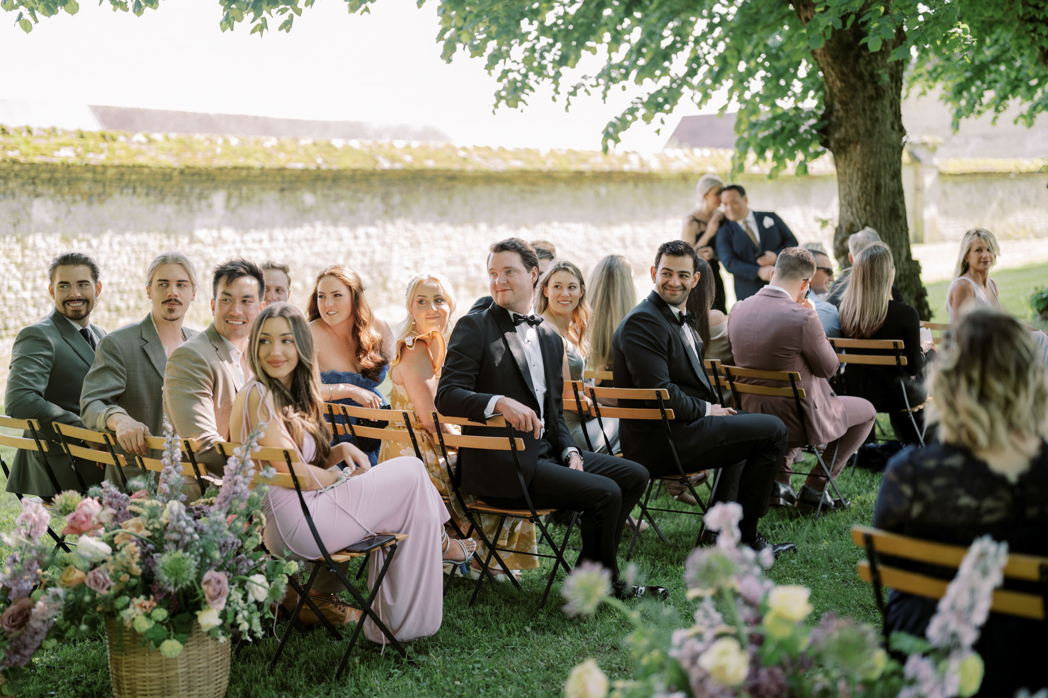 An outdoor wedding ceremony in progress, with approximately 25–30 guests seated on wooden folding chairs arranged on a lawn. Guests are dressed in a mix of formalwear including black tuxedos with bow ties, olive and tan suits, and guest attire in blush pink, dusty blue, mustard yellow, and mauve tones. Two men in black tuxedos seated centrally appear to be groomsmen. Aisle arrangements in wicker baskets filled with soft blush roses, lavender, mauve blooms, peach ranunculus, and trailing greenery line both sides of the seating area. The setting is an outdoor garden or estate grounds bordered by a stone wall, with a large tree providing partial shade over the right side of the seating. The shot is taken from a medium-wide angle at guest level, looking toward the front of the ceremony, capturing guests' relaxed, attentive expressions.