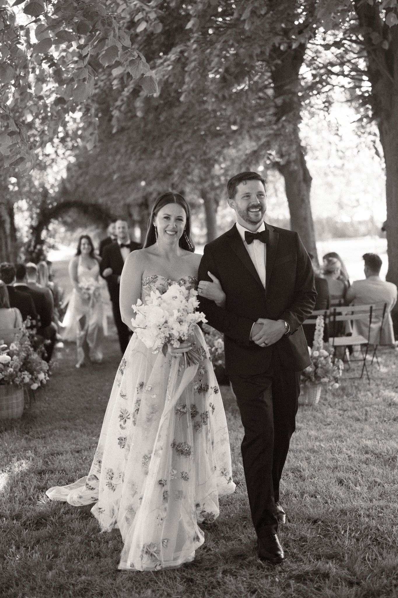 Bride in embroidered tulle gown and groom recessional under tree canopy with basket florals in B&W